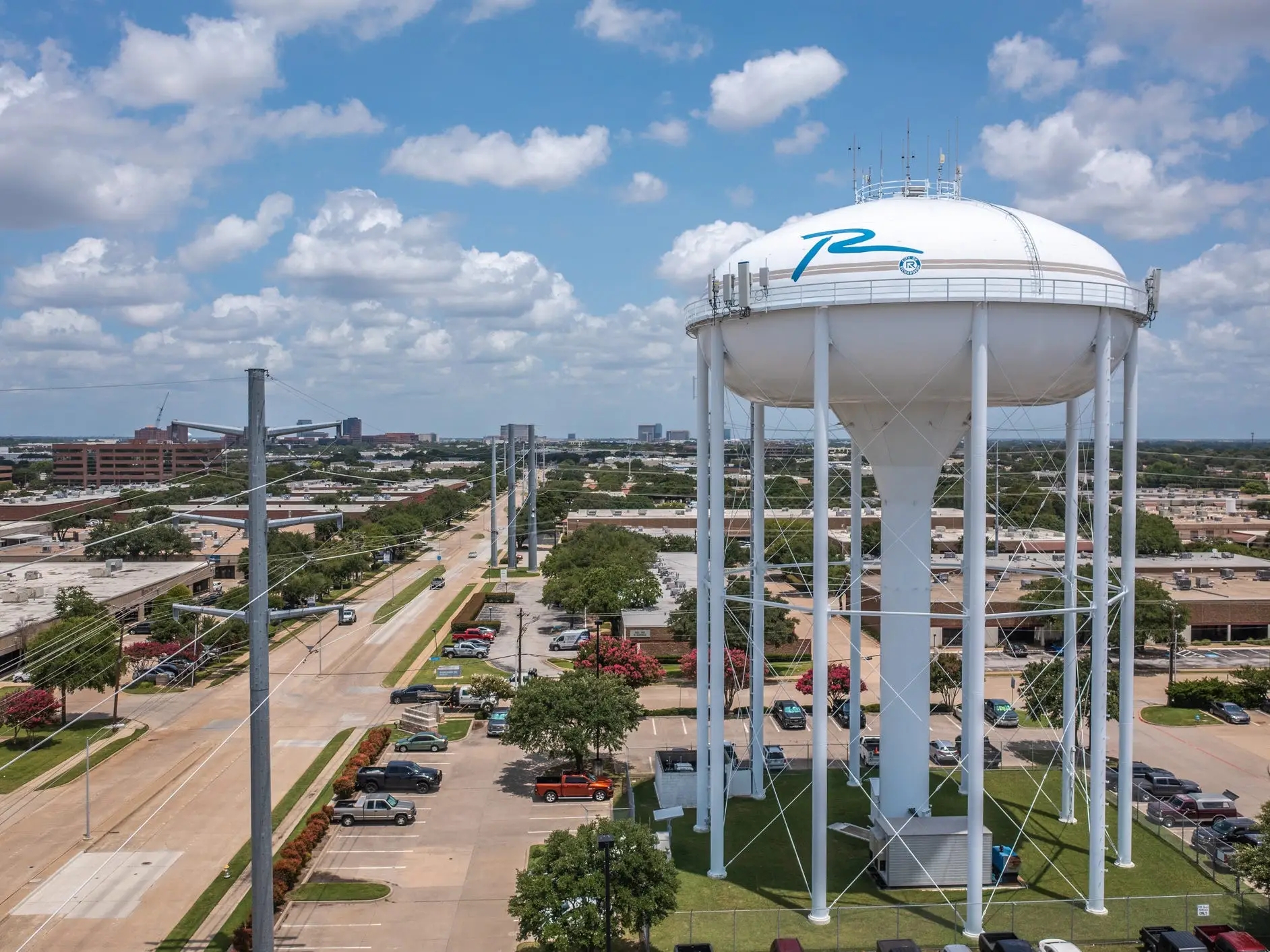 Aerial Drone View of Tall White Water Tower in Parking Lot With Parked Cars Along Street With Trees in Richardson, Texas