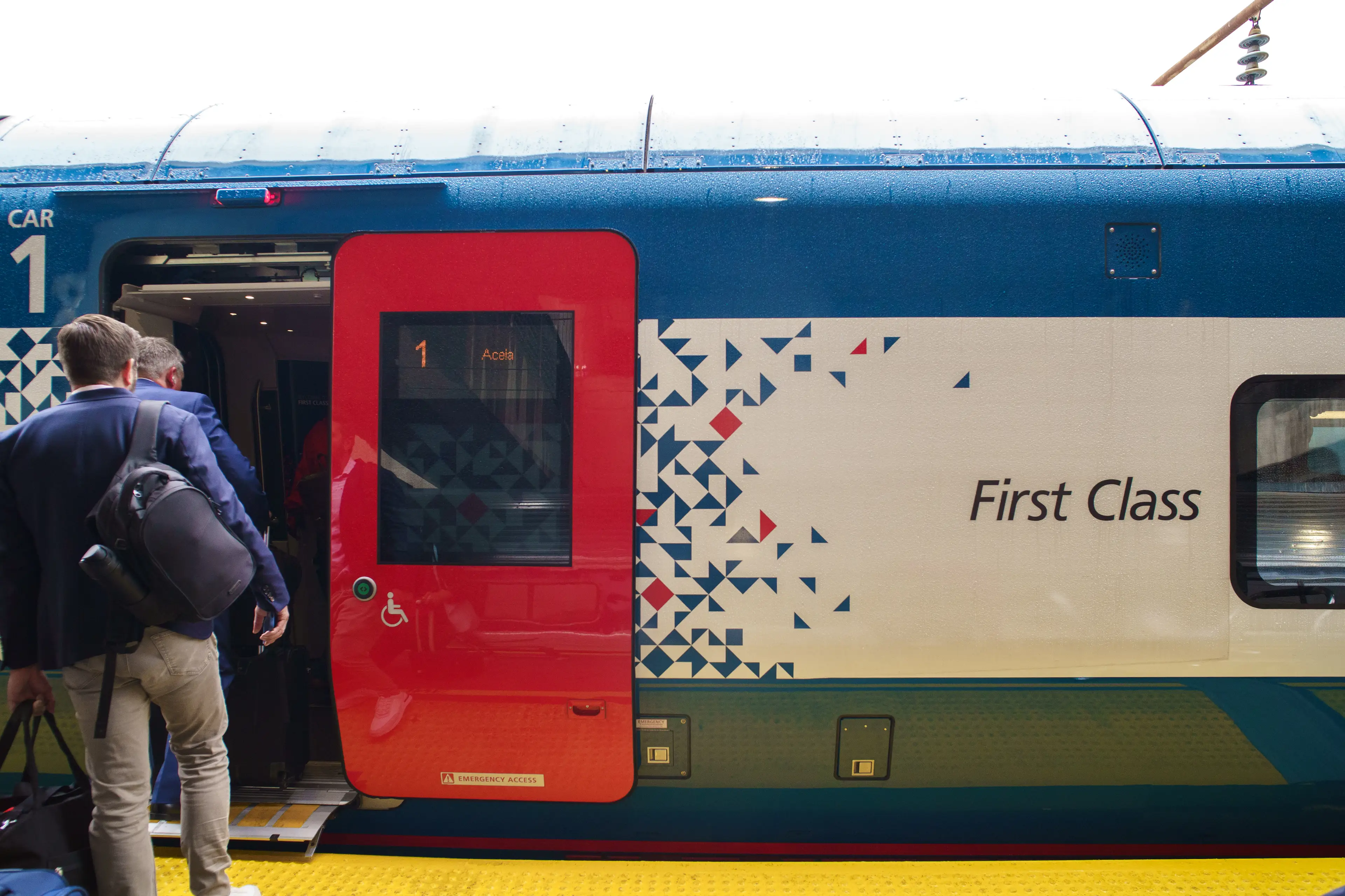 Passengers board a first-class Amtrak Acela train car with a red door at a station platform.