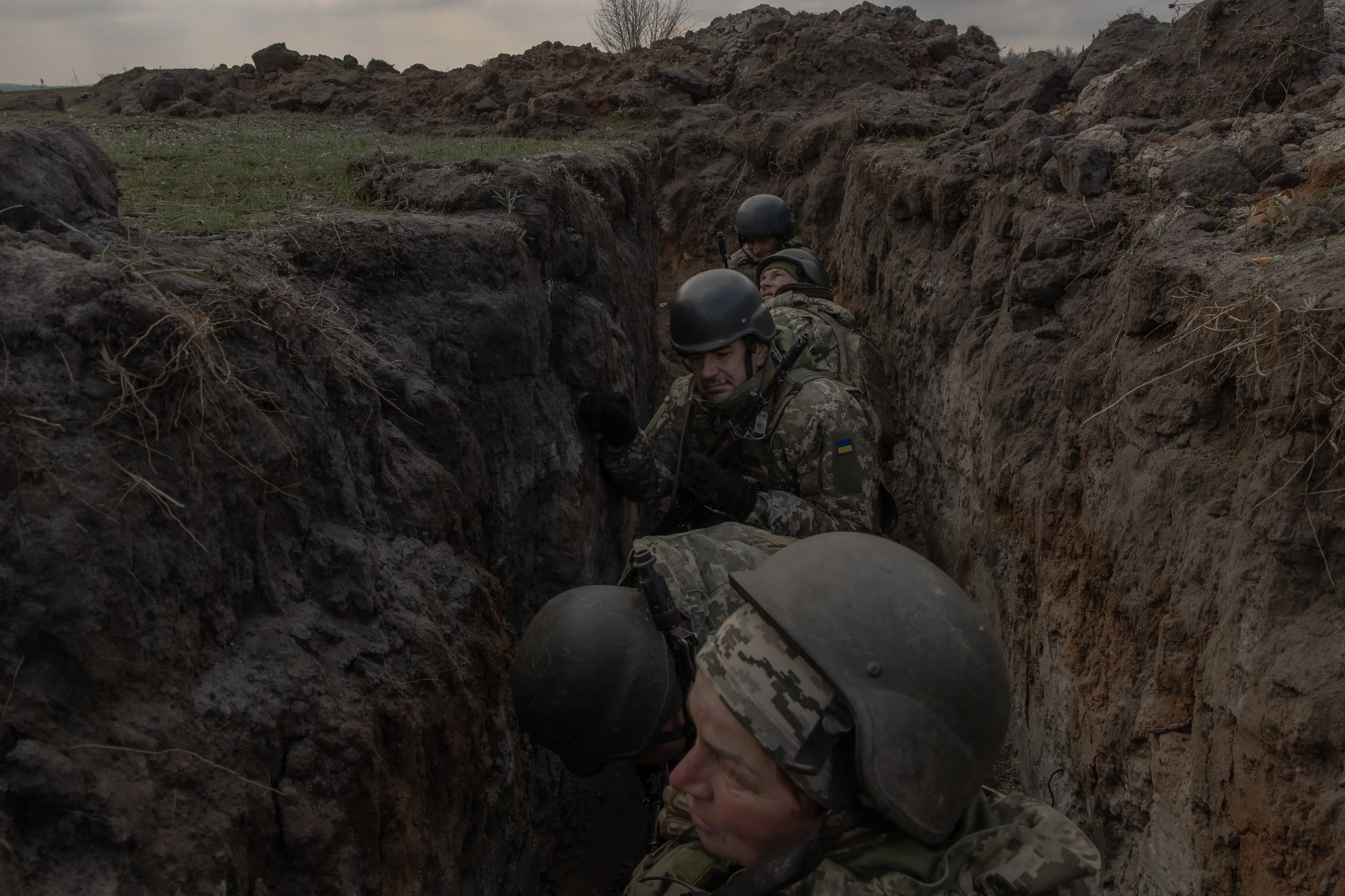 Helmeted soldiers in camouflage stand inside a muddy trench beneath an overcast sky.