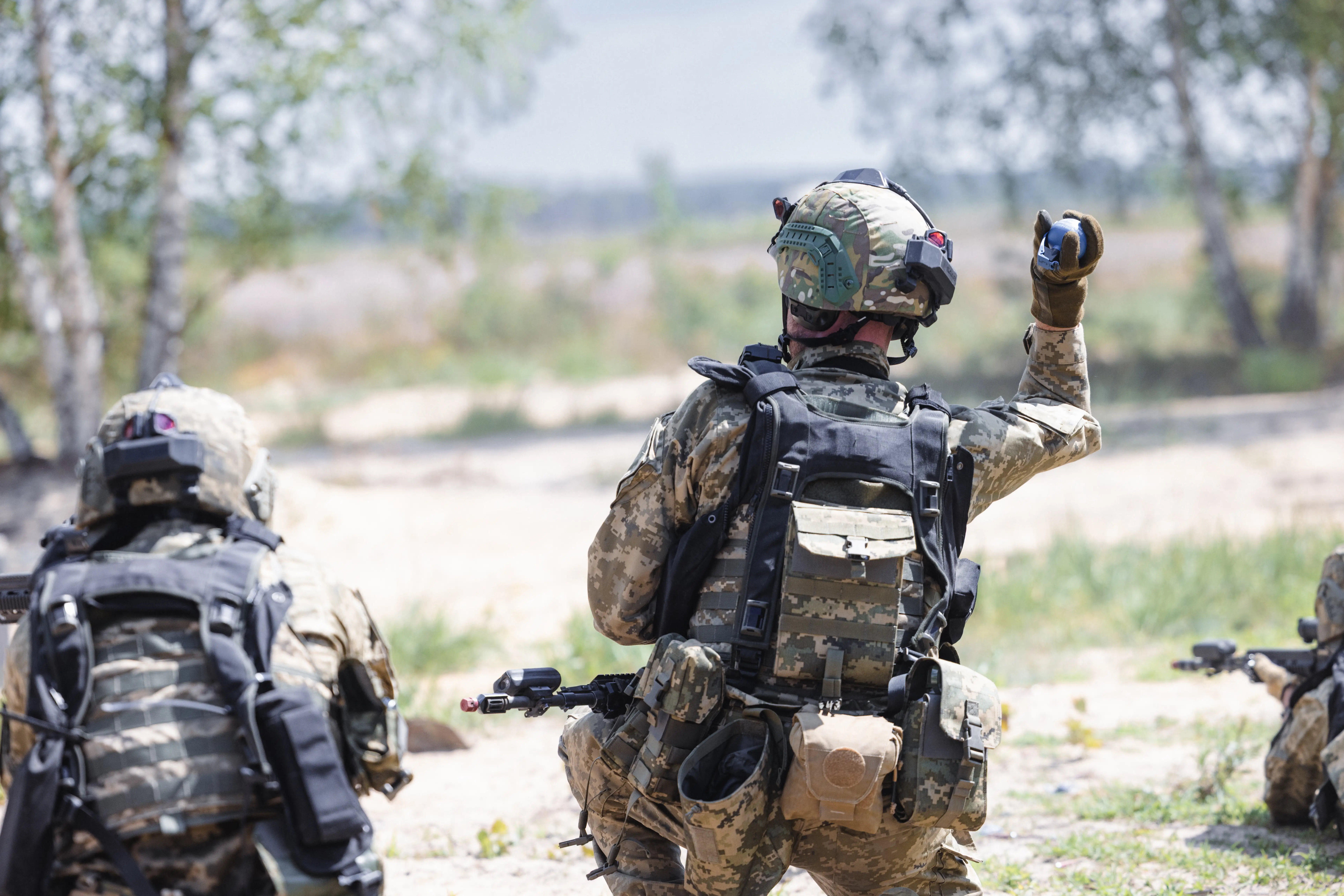Camouflaged soldiers with tactical gear train outdoors, one raising a hand signal while others crouch with rifles.