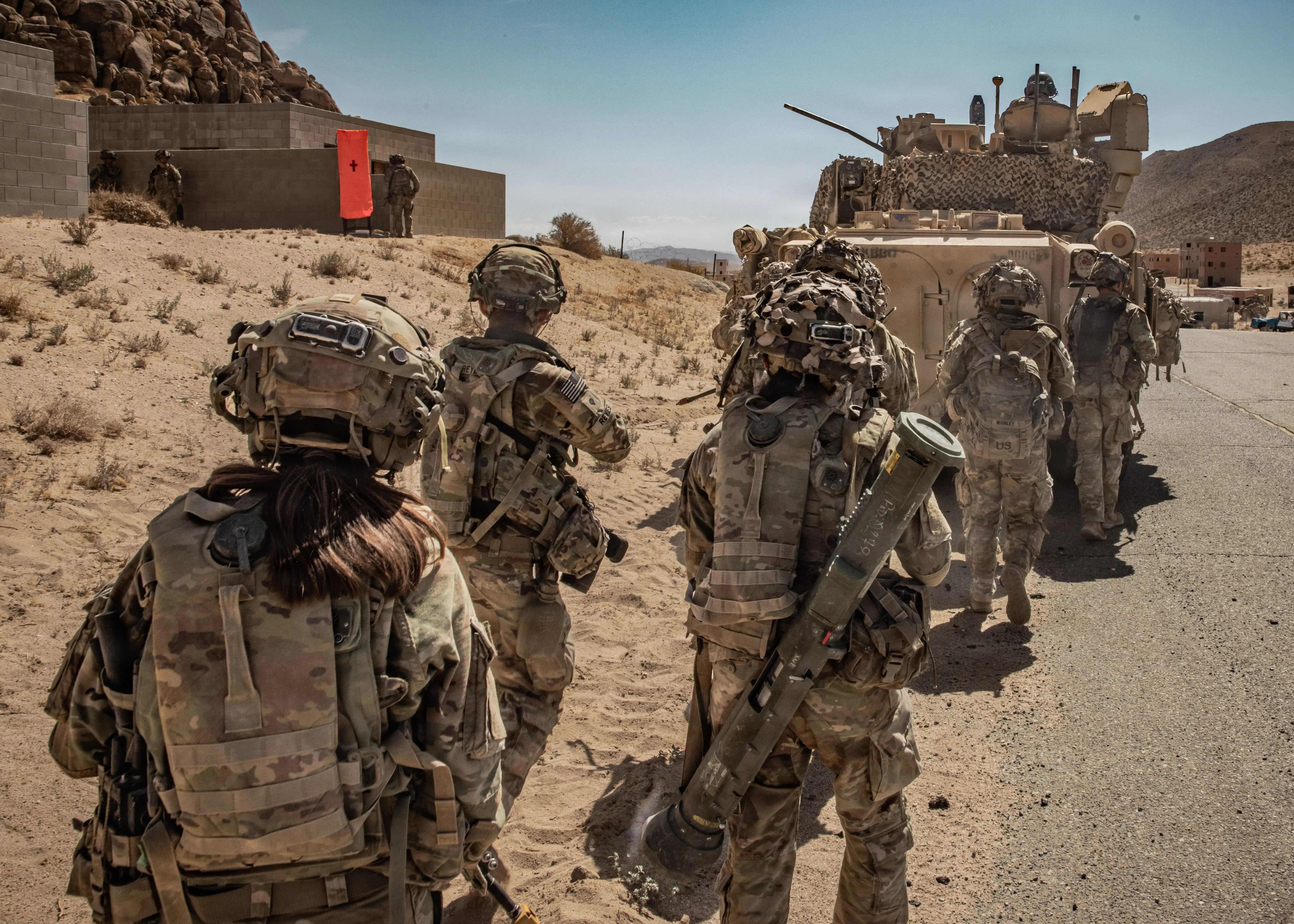 A line of US Army soldiers walks behind a Bradley Fighting Vehicle on a dirt road.