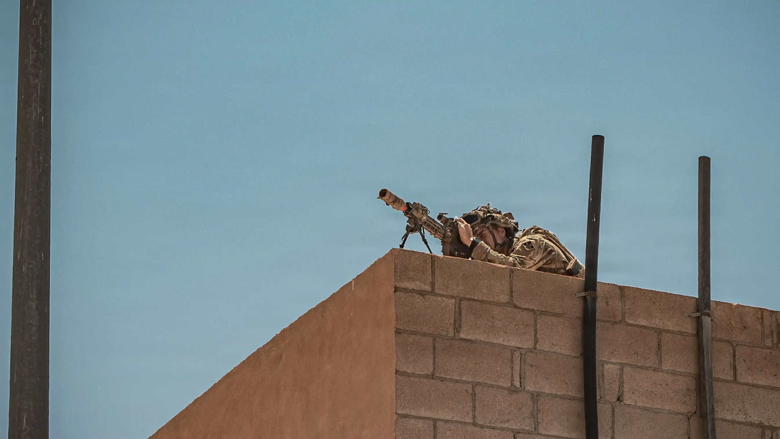 A US Army soldier crouches on a rooftop with a sniper rifle.