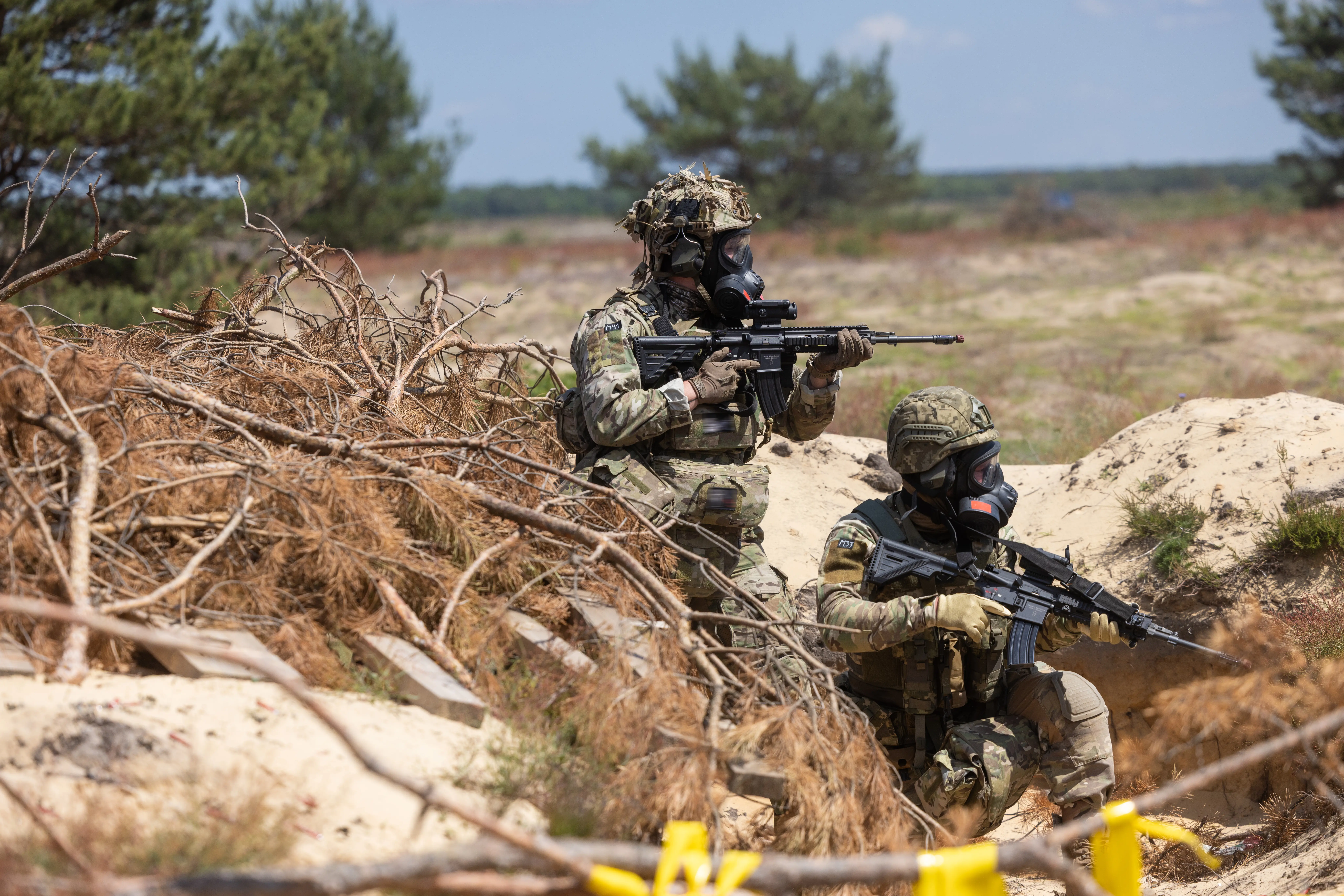 Two camouflaged soldiers wearing gas masks hold rifles in a sandy outdoor training area with brush cover.