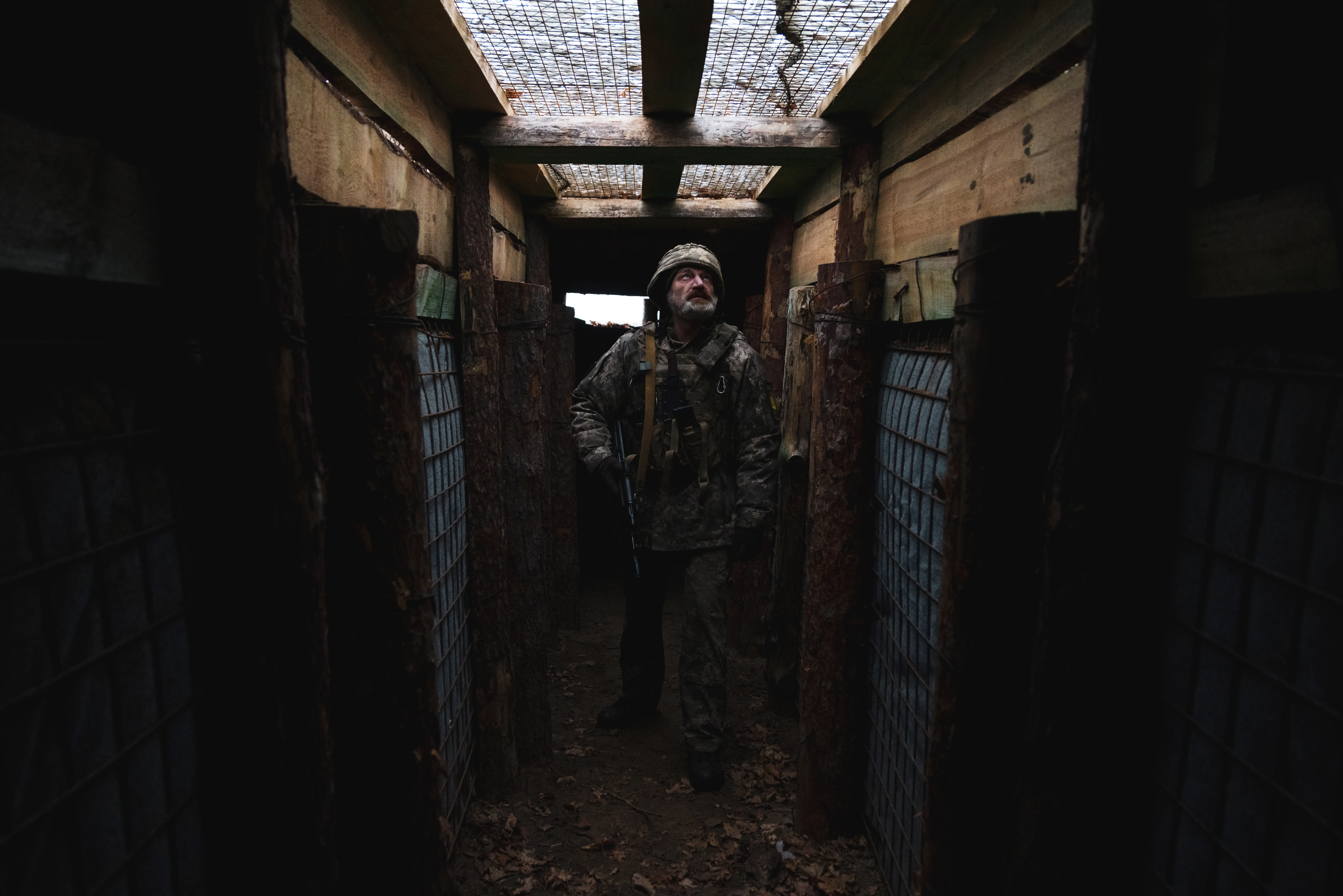 Camouflaged armed person stands in a narrow wooden trench or bunker corridor under a grated overhead opening.
