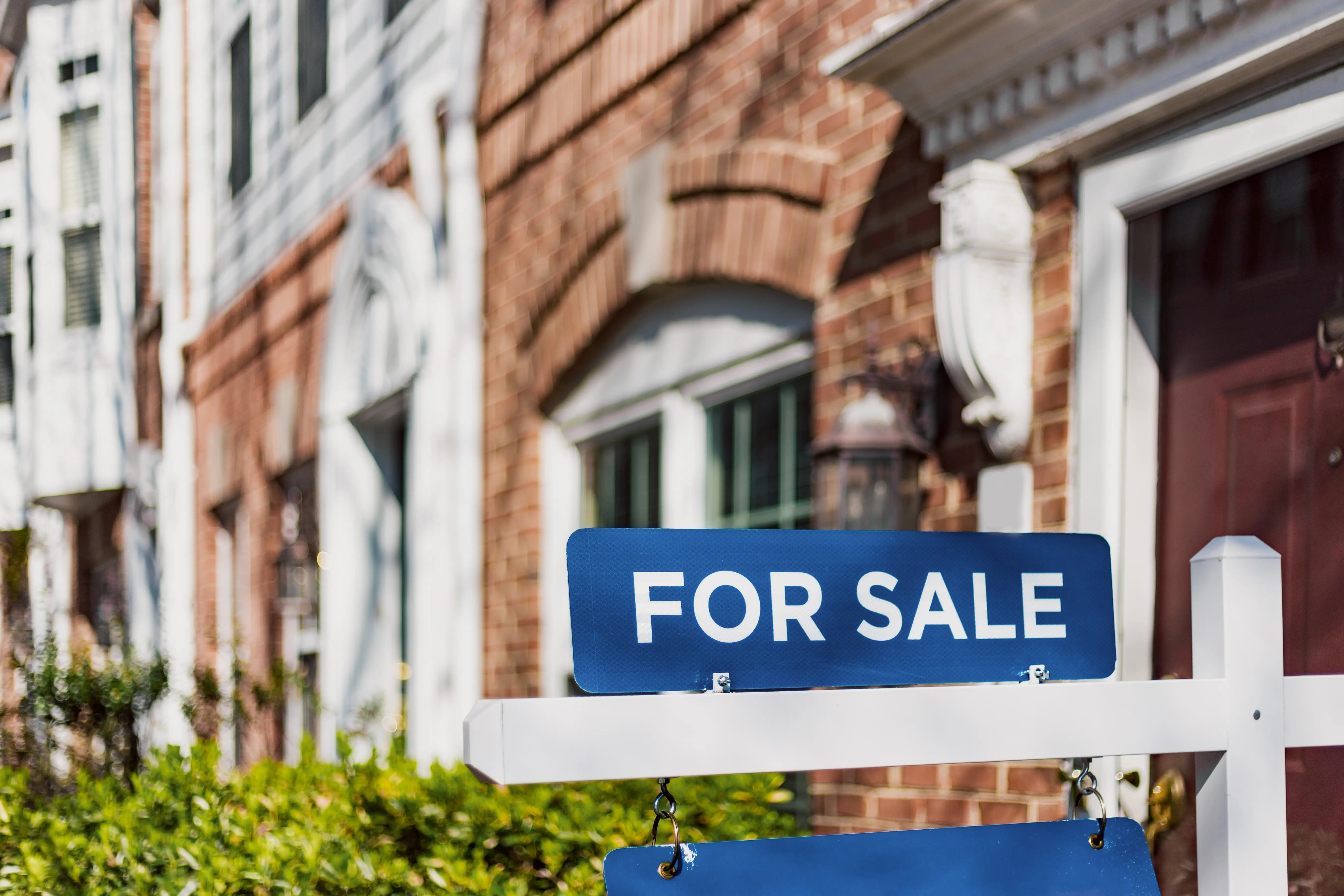 Blue “FOR SALE” sign stands in front of brick row houses on a residential street.
