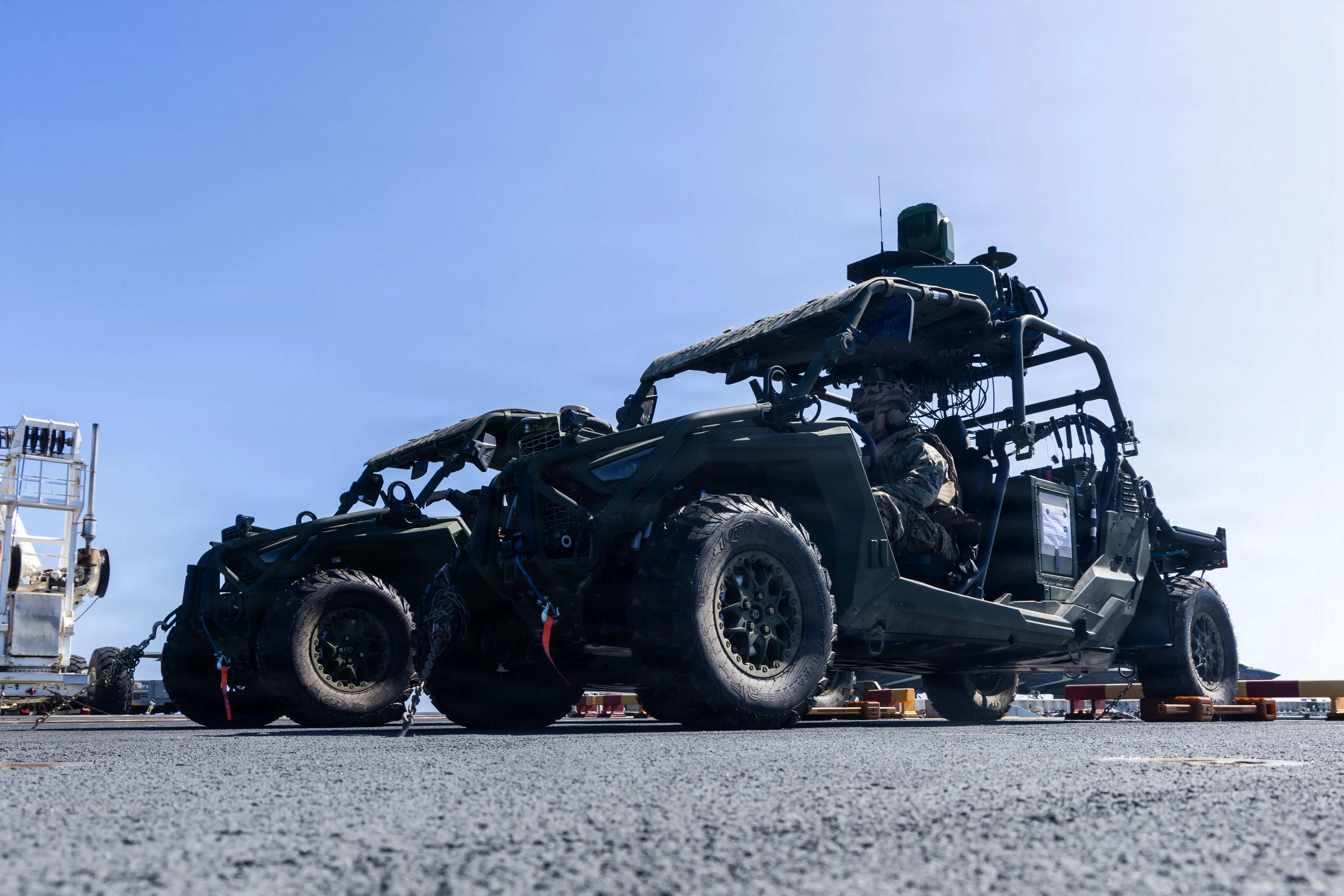 Military all-terrain vehicles are secured on a deck under a clear blue sky.