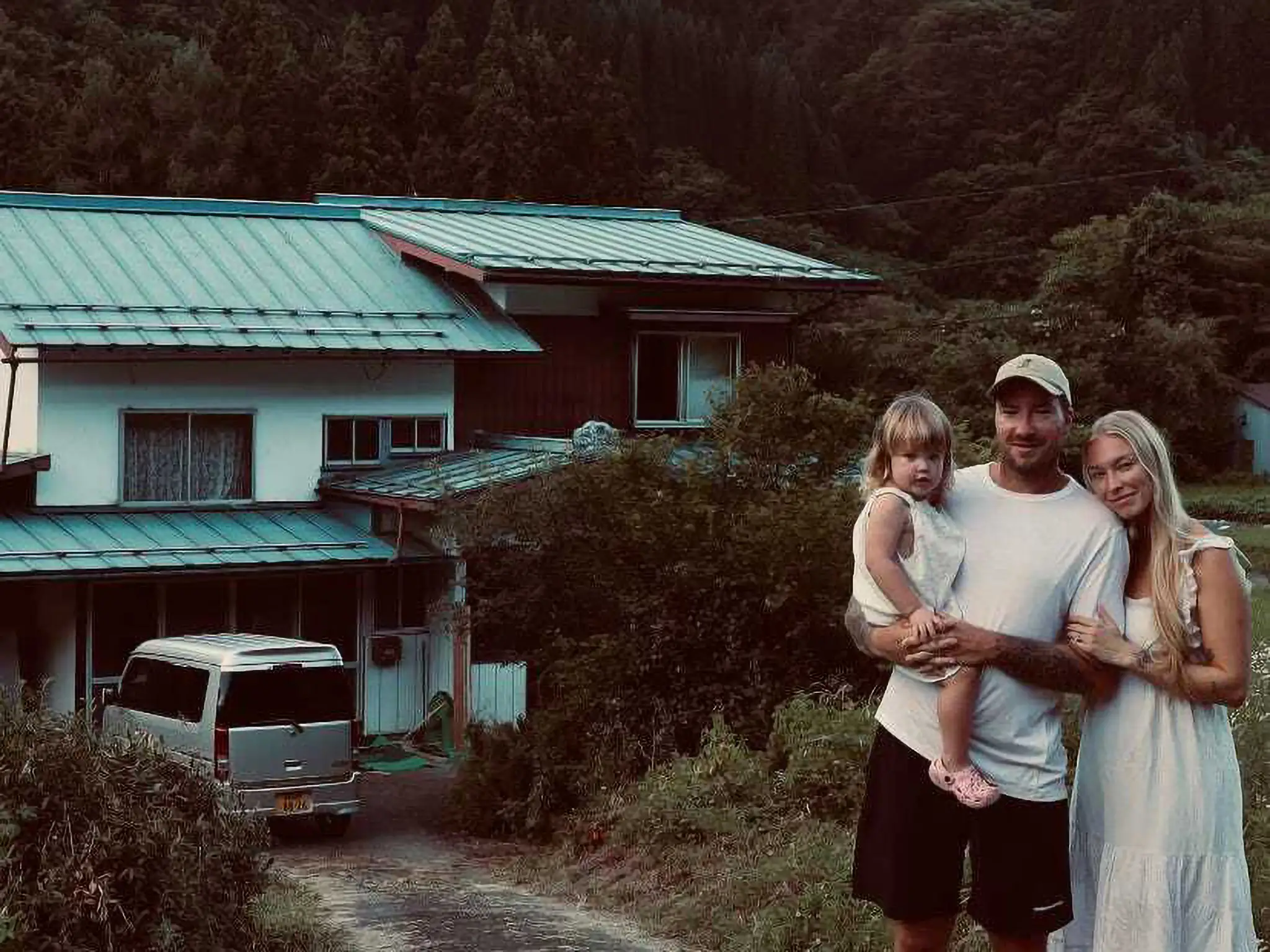 A couple stands in front of a house holding their daughter.