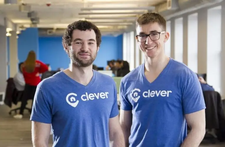 Two people wearing blue Clever T-shirts stand in a bright open office workspace.