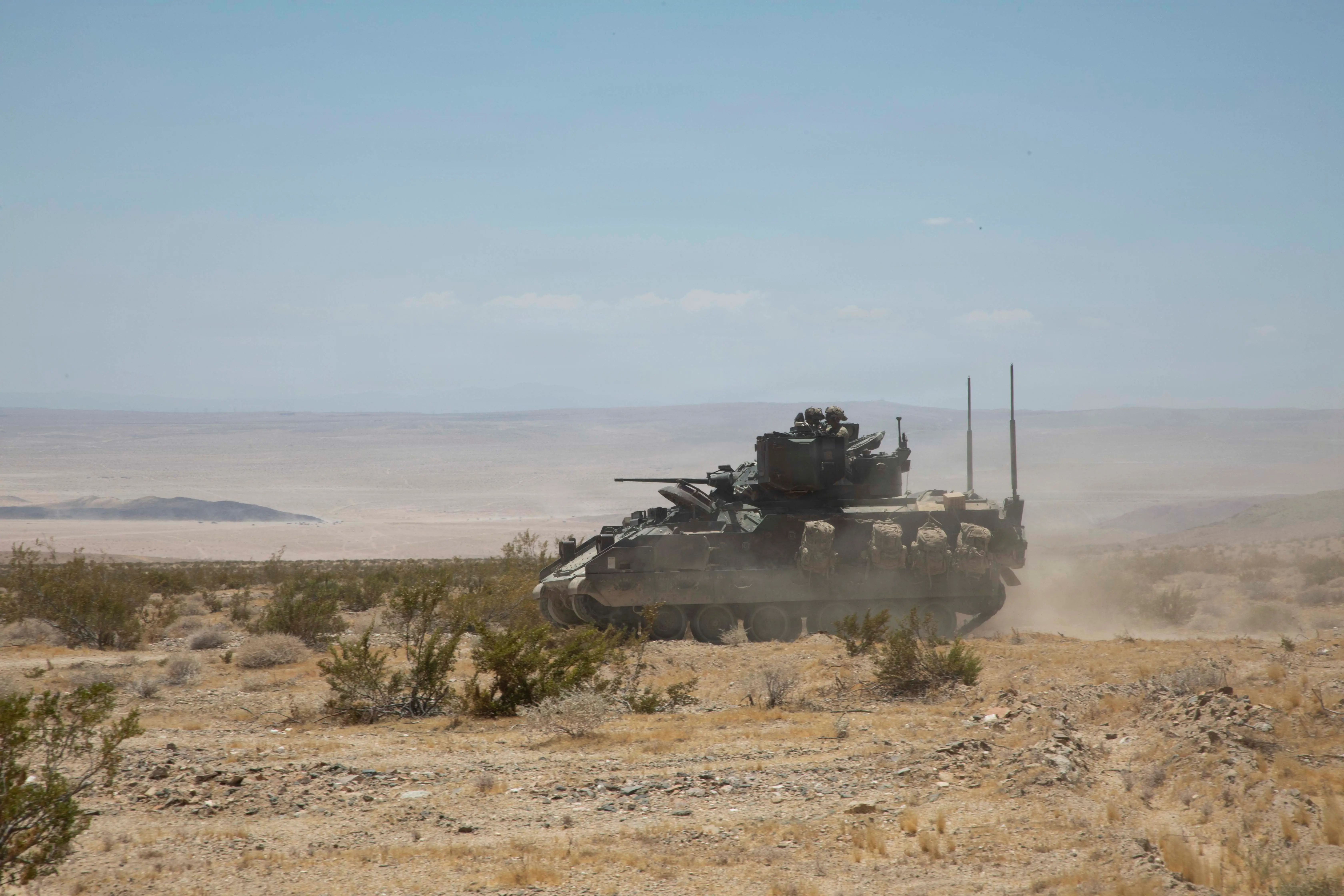 A US Army Bradley Fighting Vehicle sits on the dusty desert terrain with a blue sky in the background.