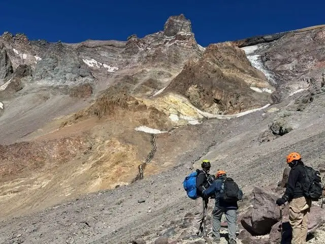 Three hikers with helmets and backpacks traverse a rocky high mountain slope beneath rugged peaks and patches of snow.