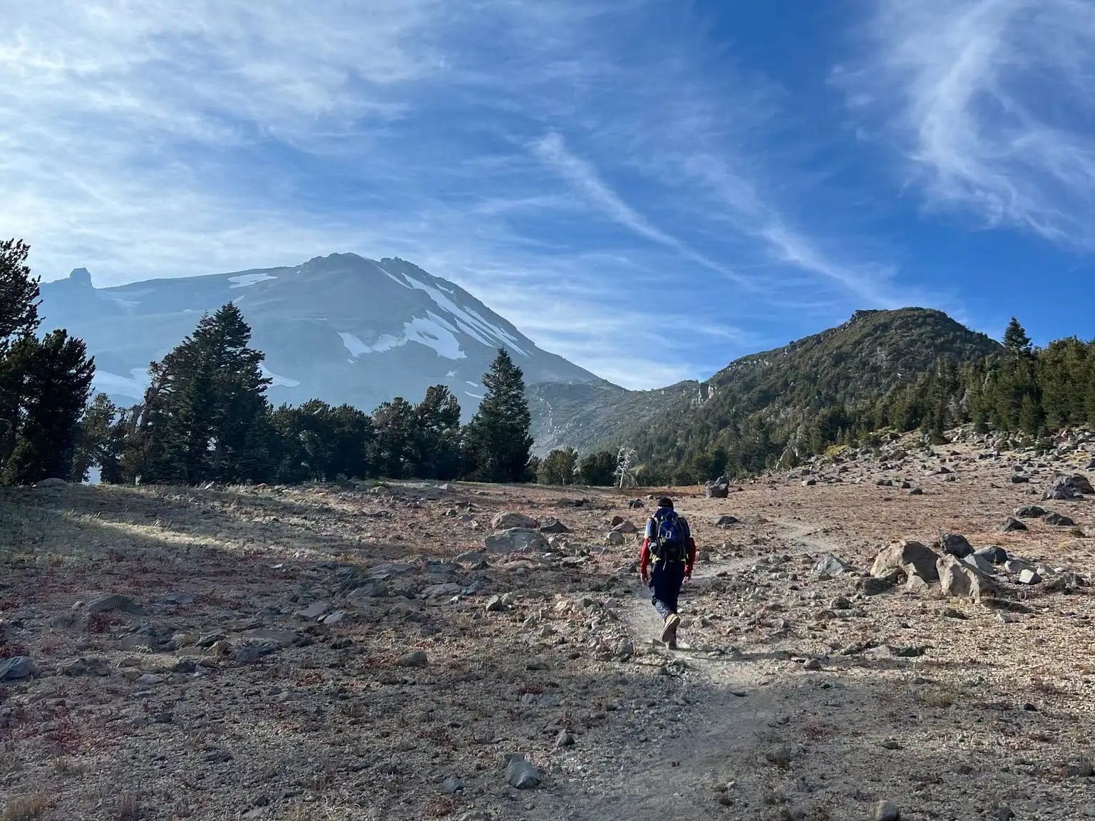 A hiker walks along a rocky mountain trail beneath a blue sky with wispy clouds.