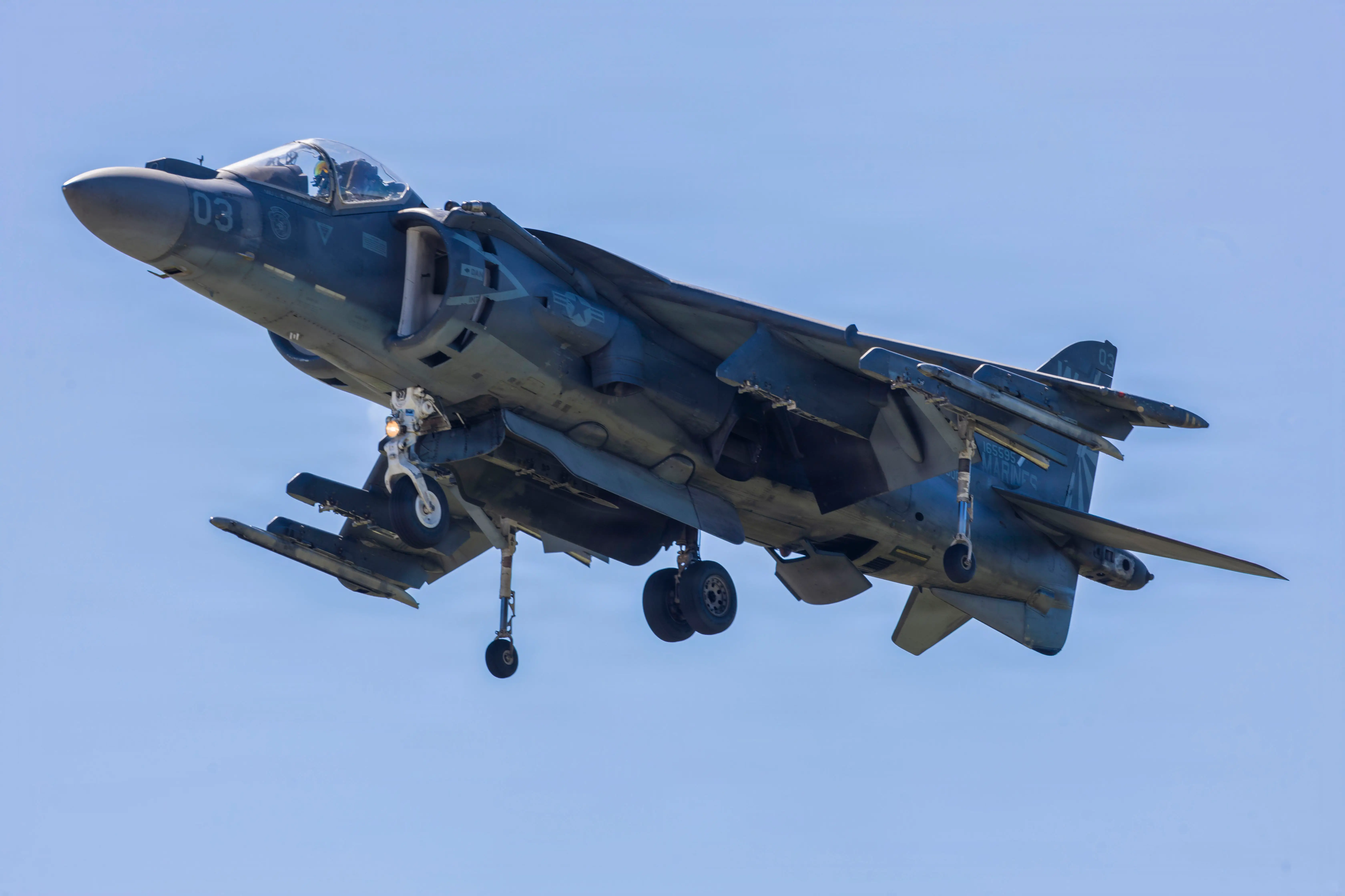 An AV-8B Harrier jet flies with landing gear down against a clear blue sky.