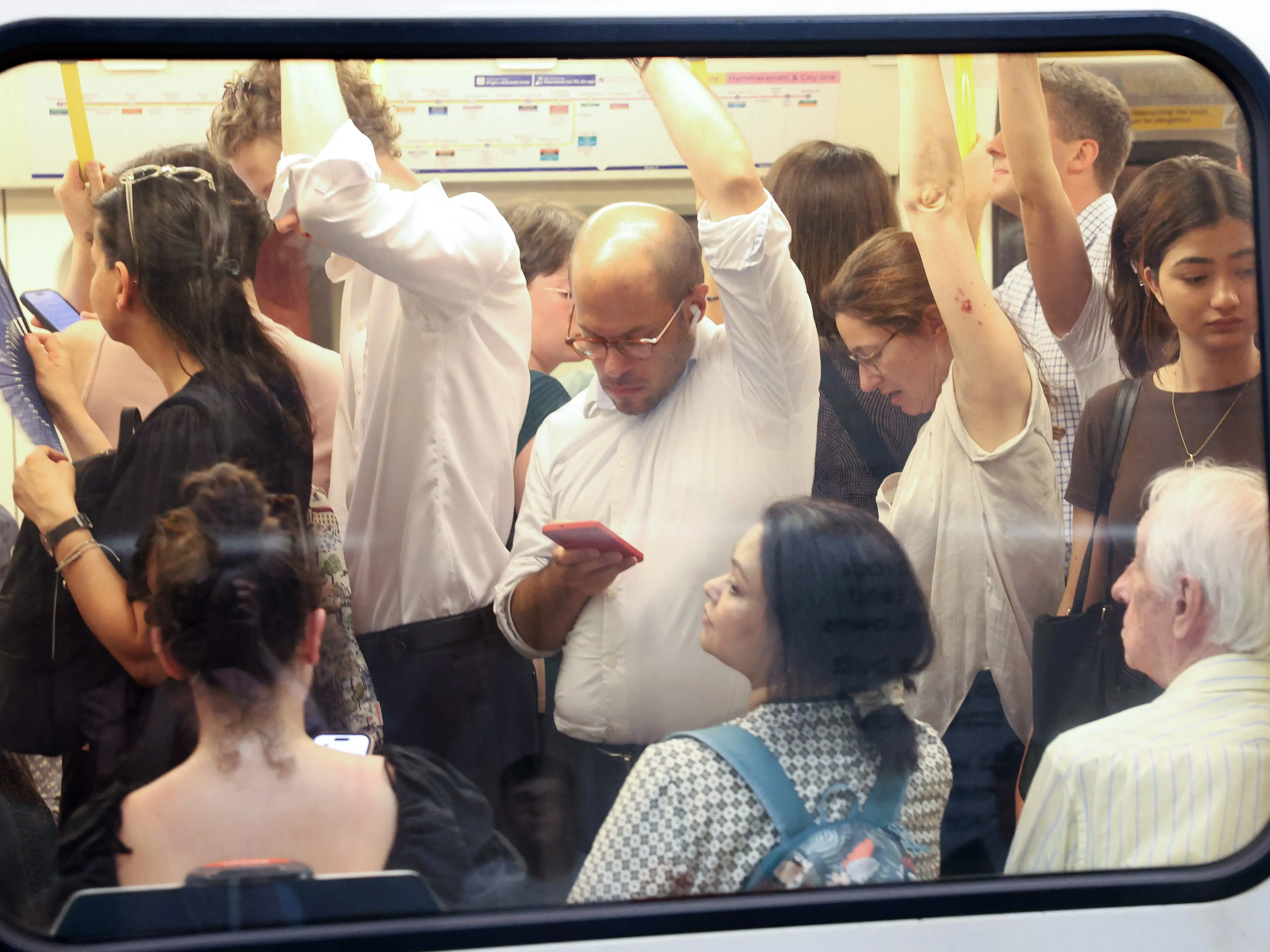 Crowded public transit car seen through a window, with passengers standing, seated, holding rails, and using smartphones.