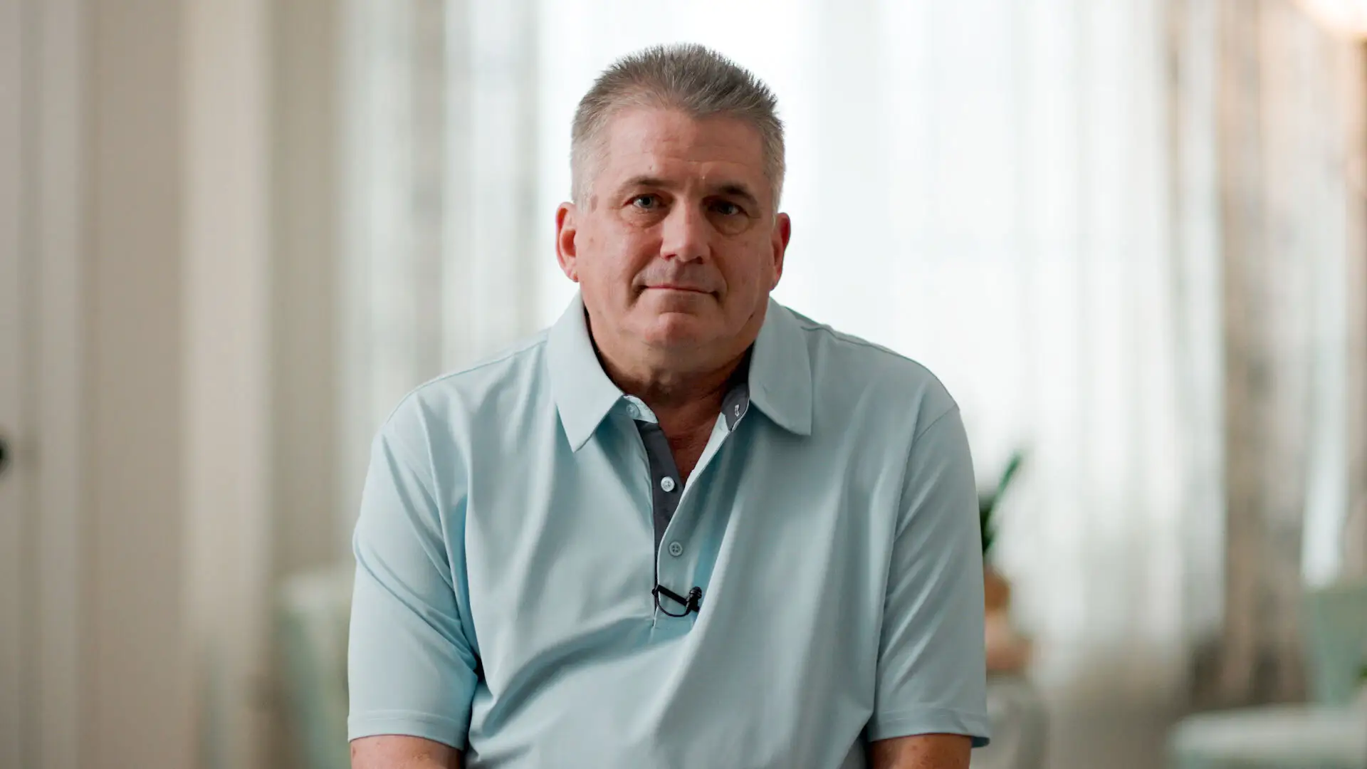 Bill Lewis in a blue polo sitting in a lit room with white sheer curtains.