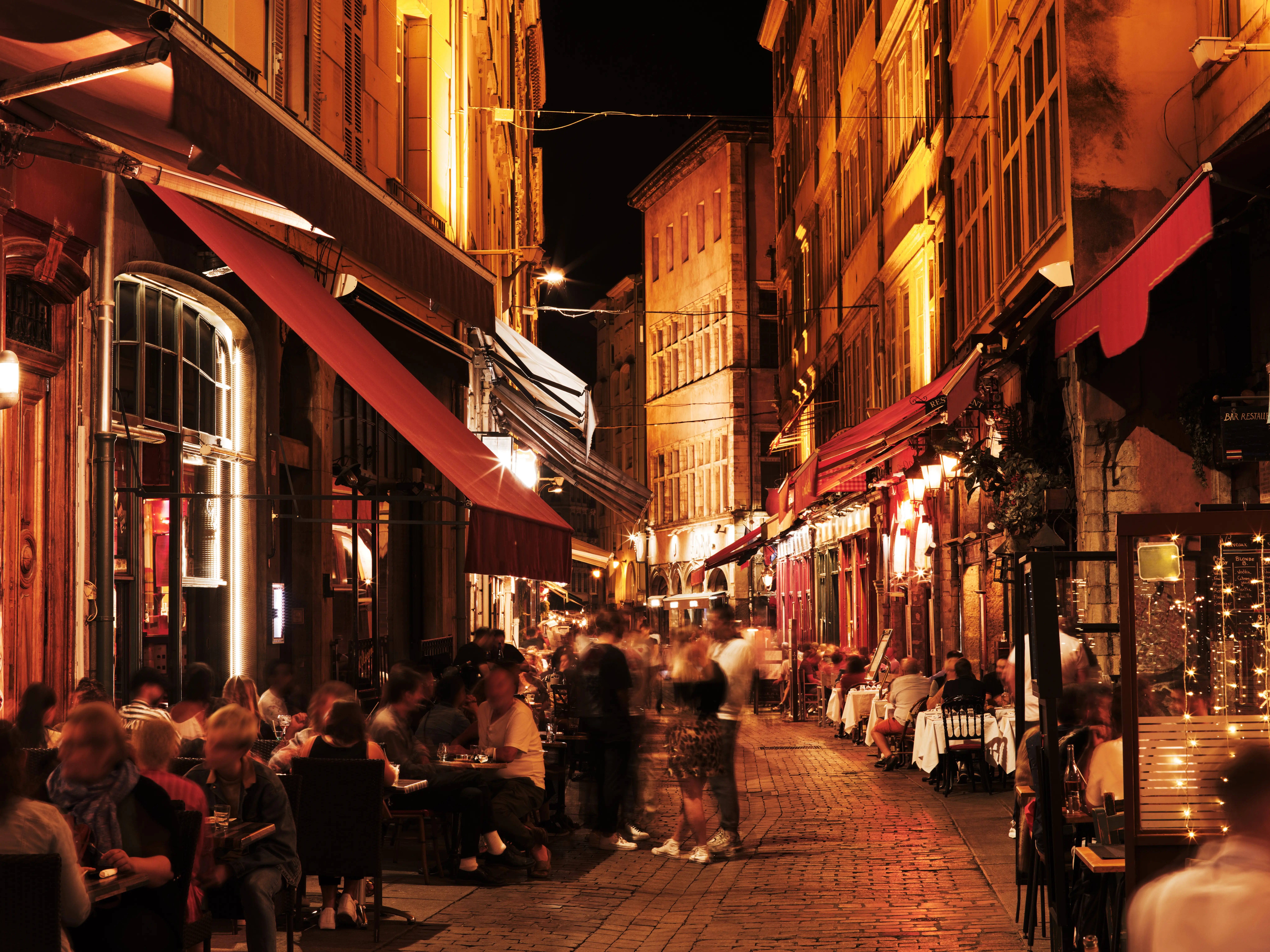 Warmly lit narrow cobblestone street with outdoor restaurant seating and blurred diners at night.