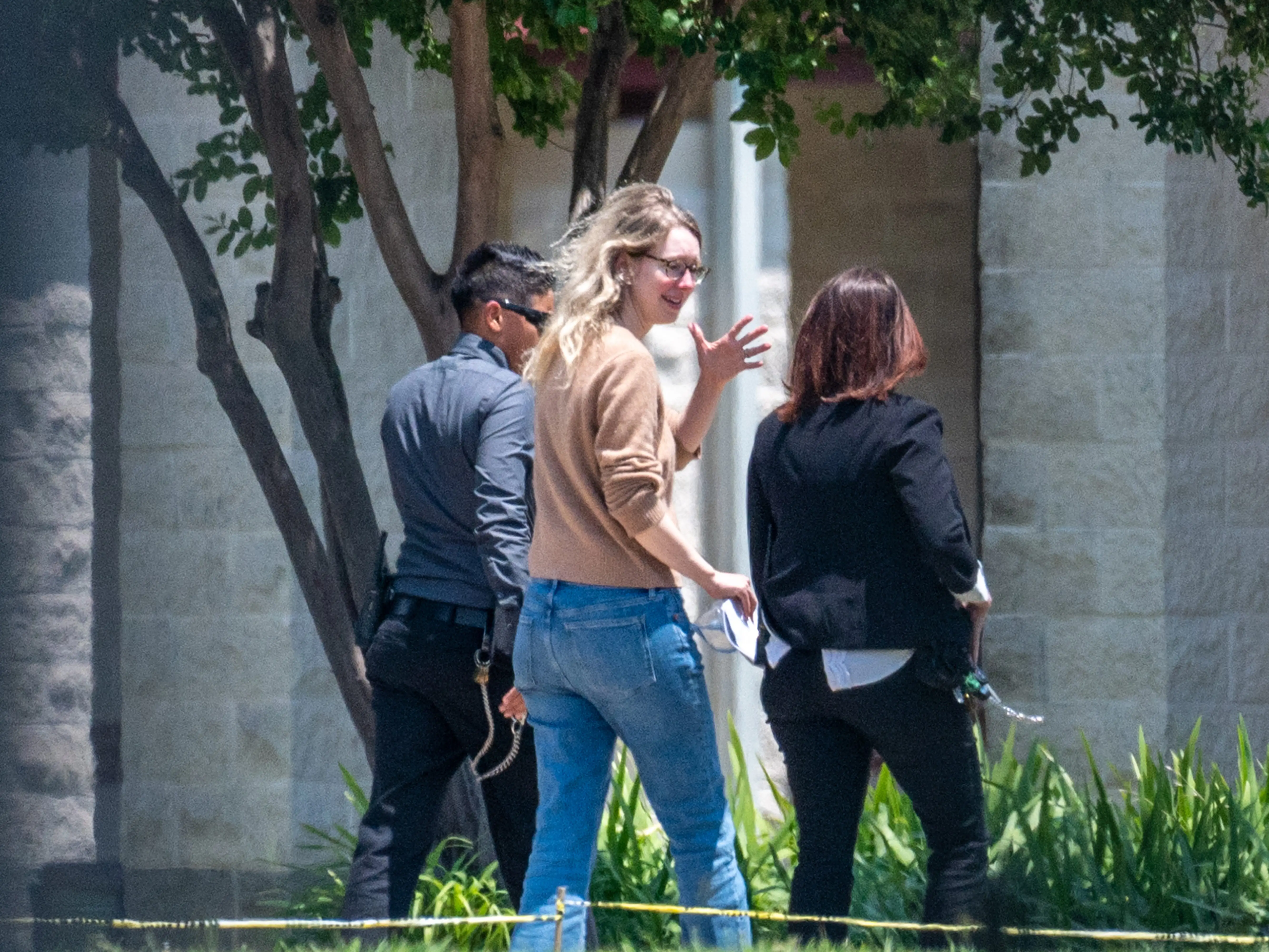 Elizabeth Holmes waves while walking with two others past trees and landscaping outside a stone building.