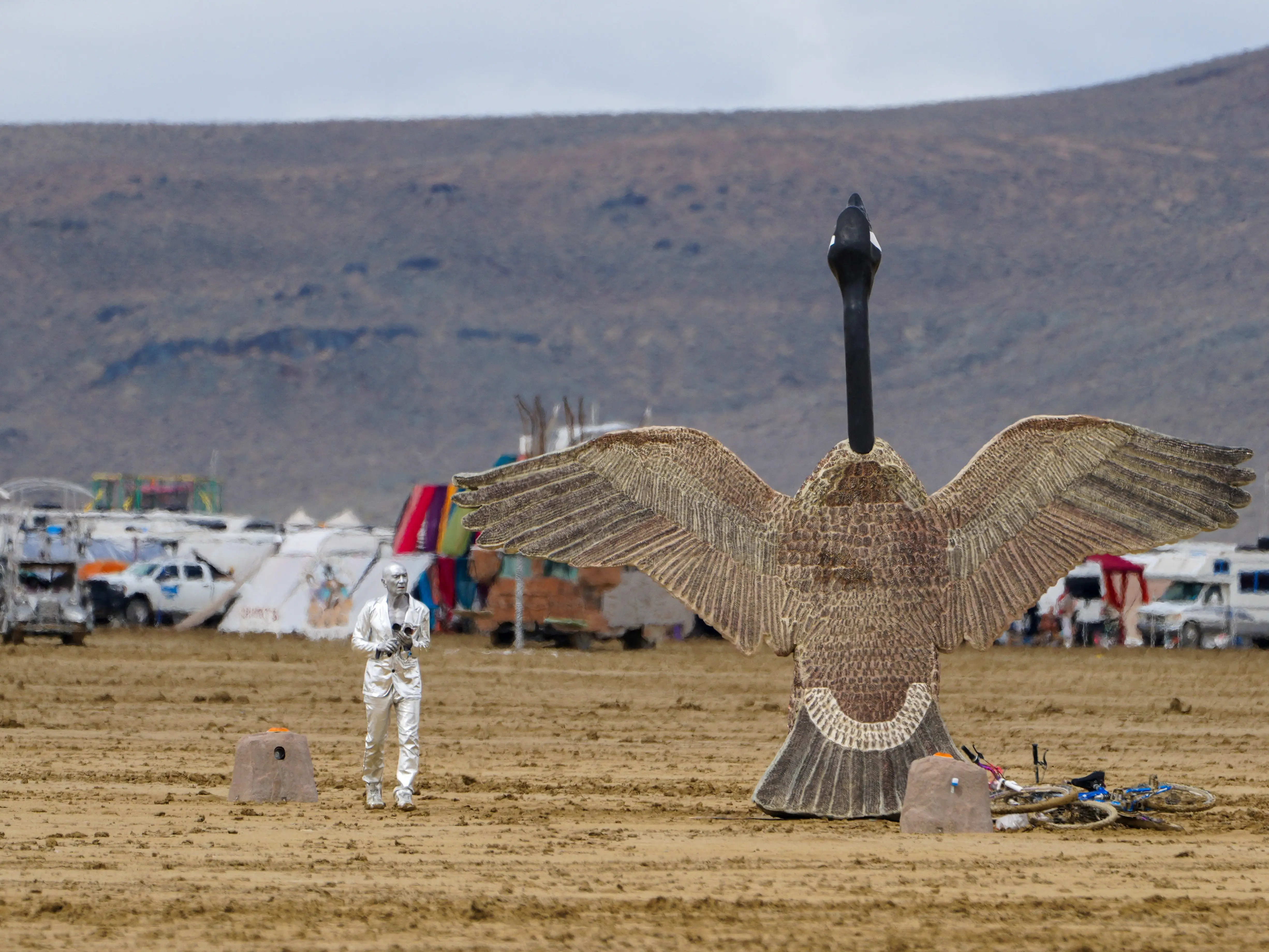 A large bird sculpture stands on a desert plain near a silver-clad person, tents, vehicles, and mountains.