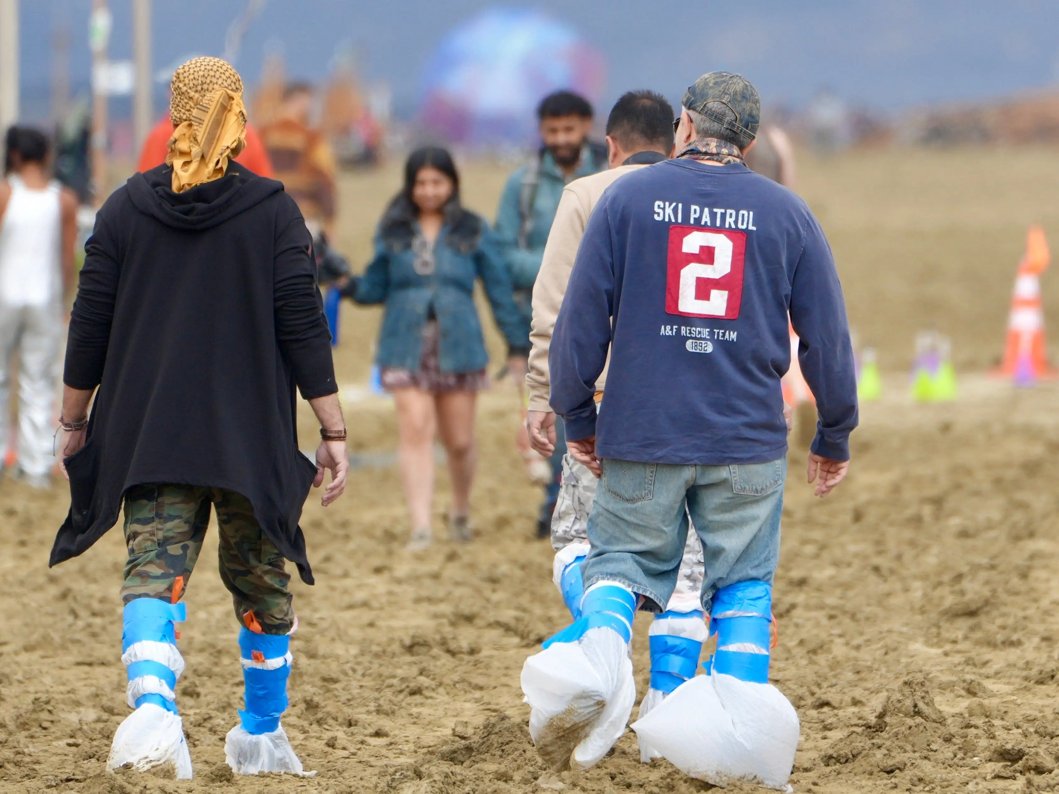 People walk across a muddy outdoor site wearing blue-and-white protective shoe covers.