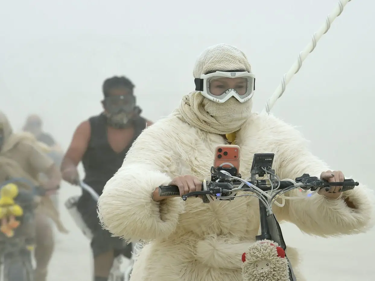 Costumed cyclists wearing goggles ride through a dusty outdoor scene.