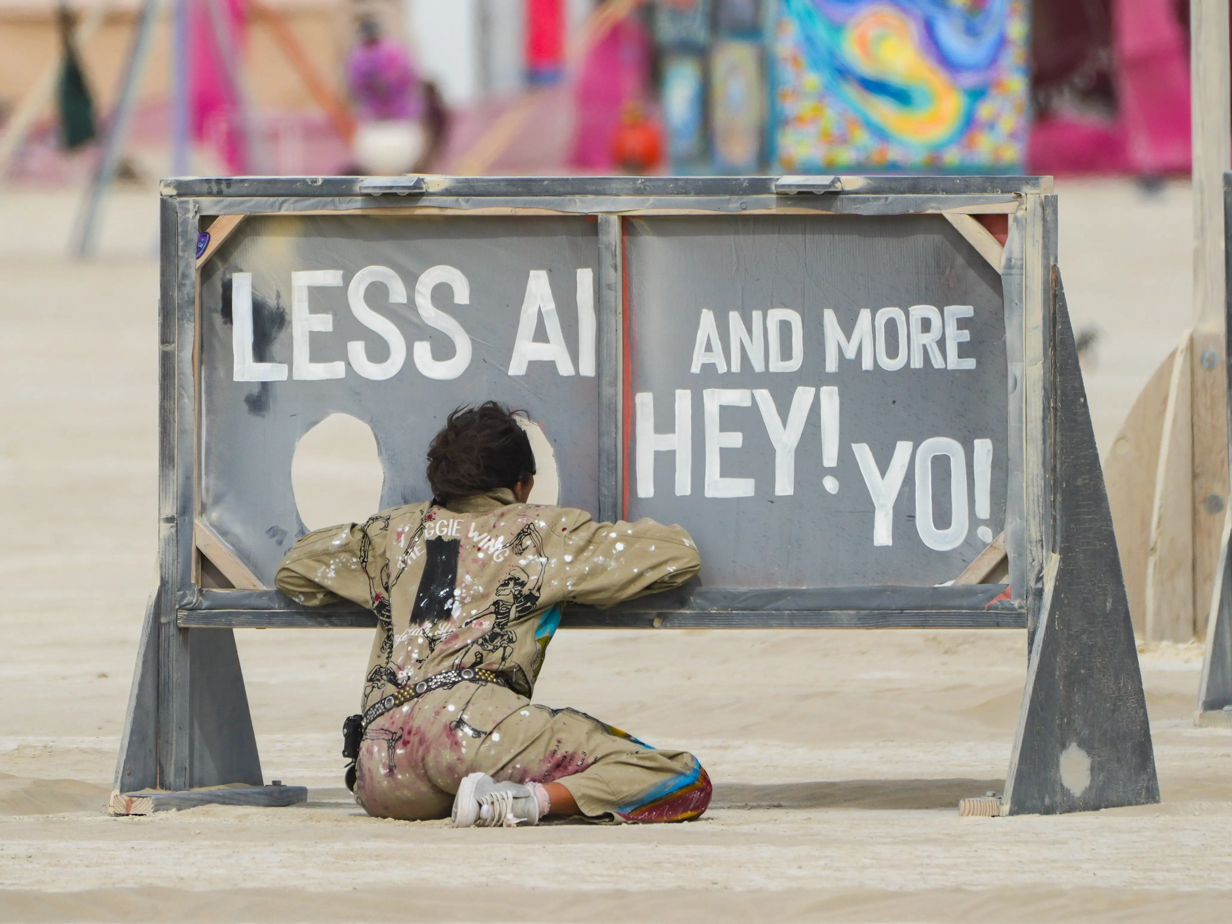 Person in paint-splattered clothing kneels beside a large painted sign with bold white lettering at an outdoor art site.