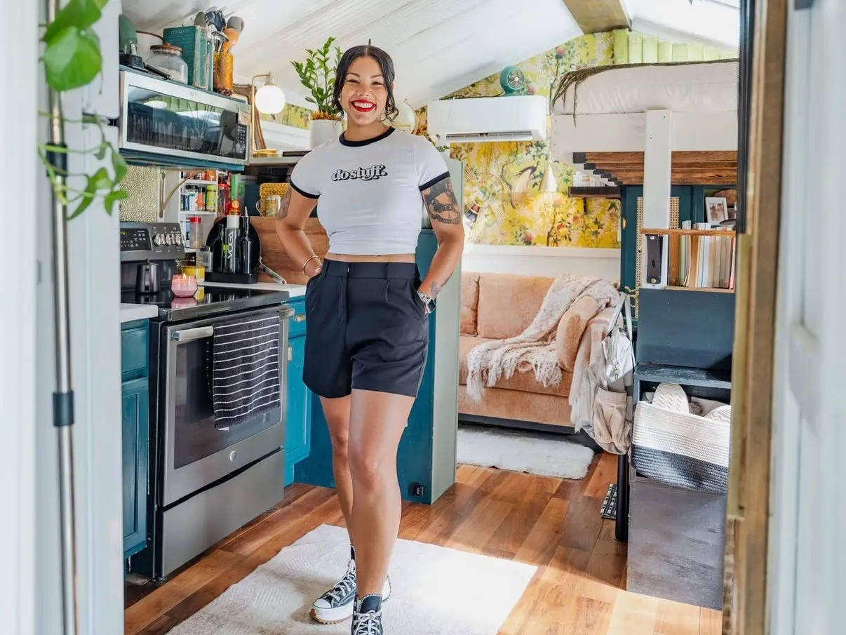 Margo Fairchild stands in a tiny home kitchen with plants, a loft bed, a couch, and wood floors in the background.