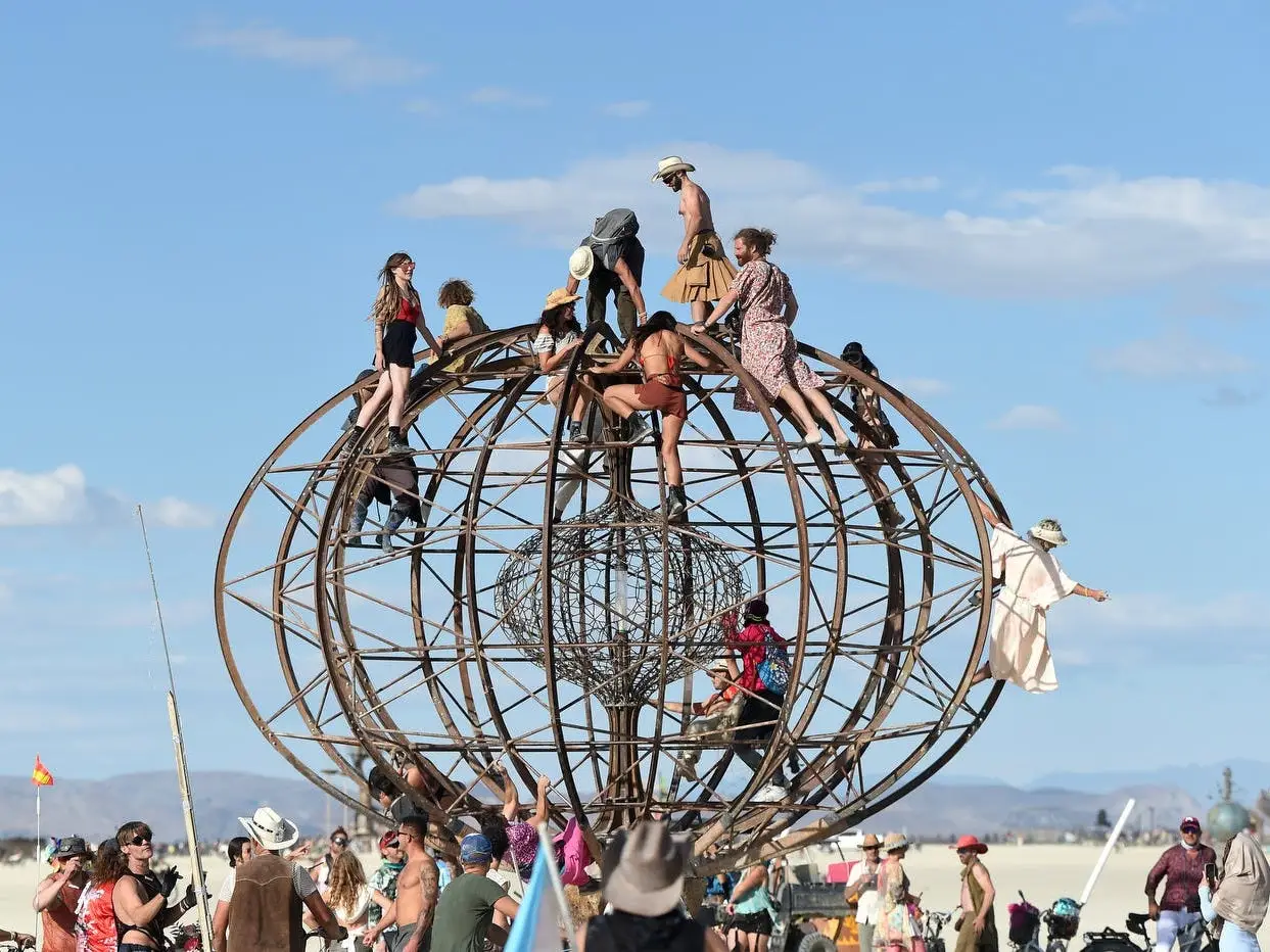 Festival attendees climb a large spherical metal structure in a desert setting under a blue sky.