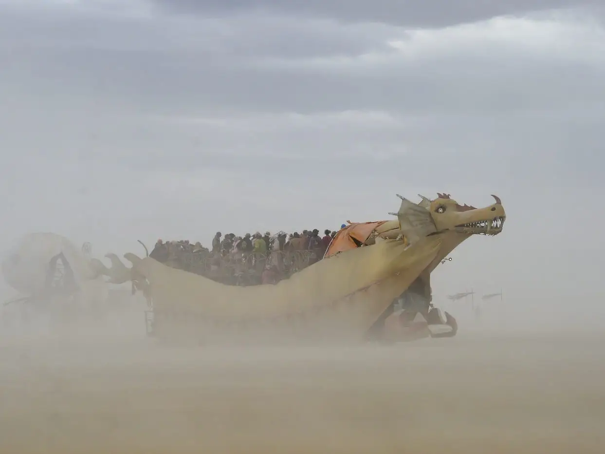Dragon-shaped art vehicle carries riders through dust under cloudy skies.