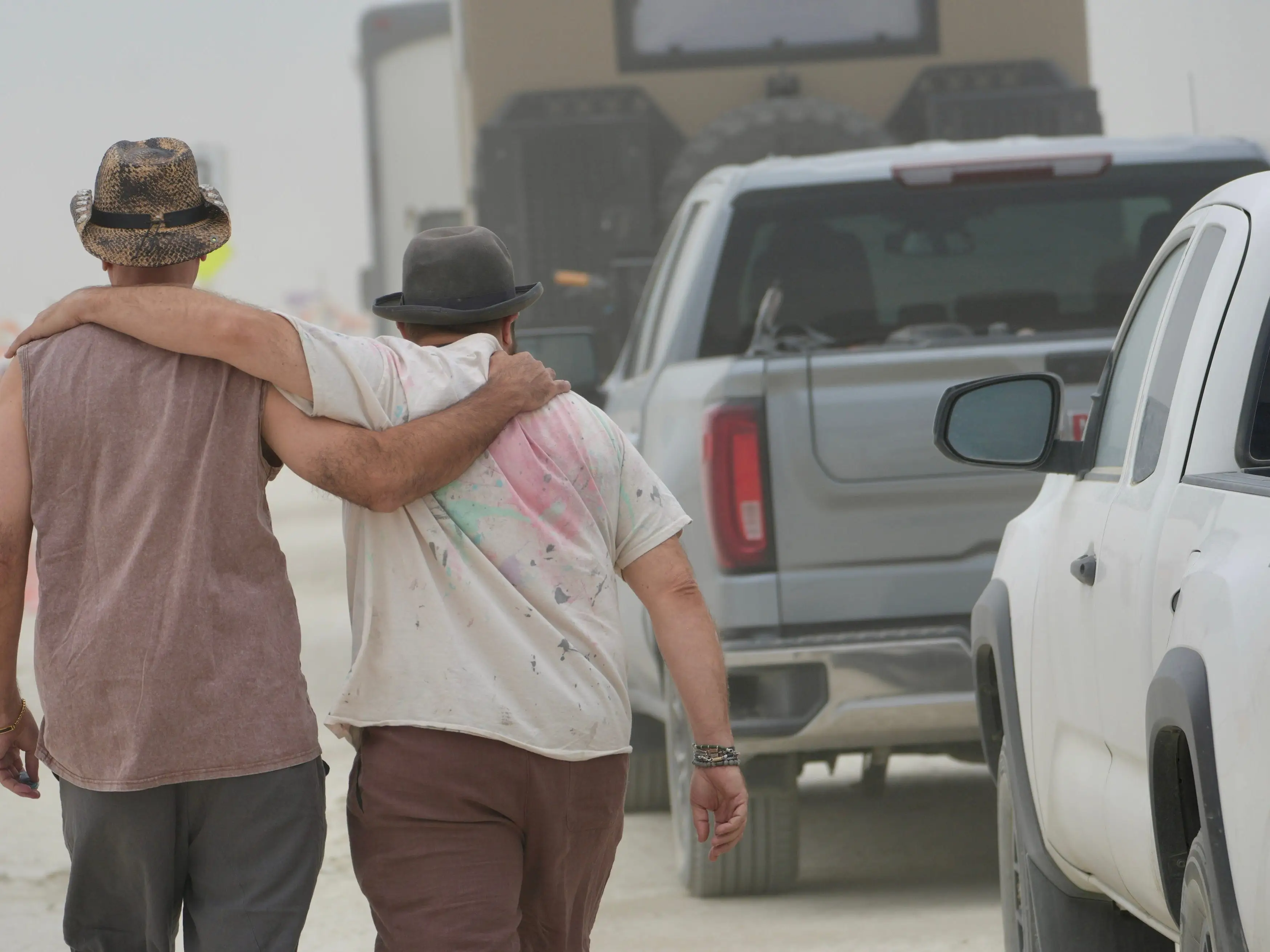 Two people walk arm in arm along a dusty road beside pickup trucks.