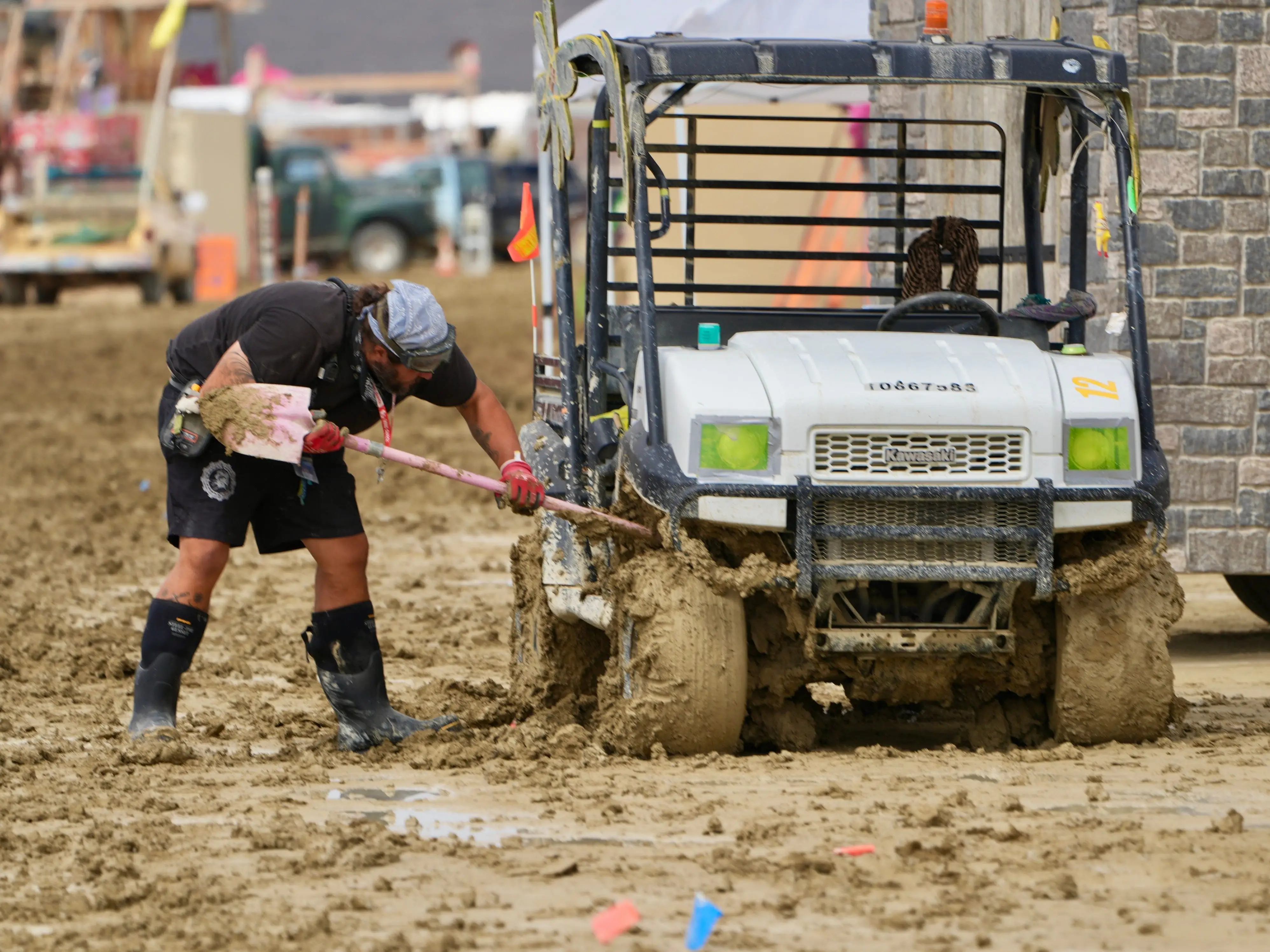 Worker shovels thick mud beside a utility vehicle stuck near a stone building.
