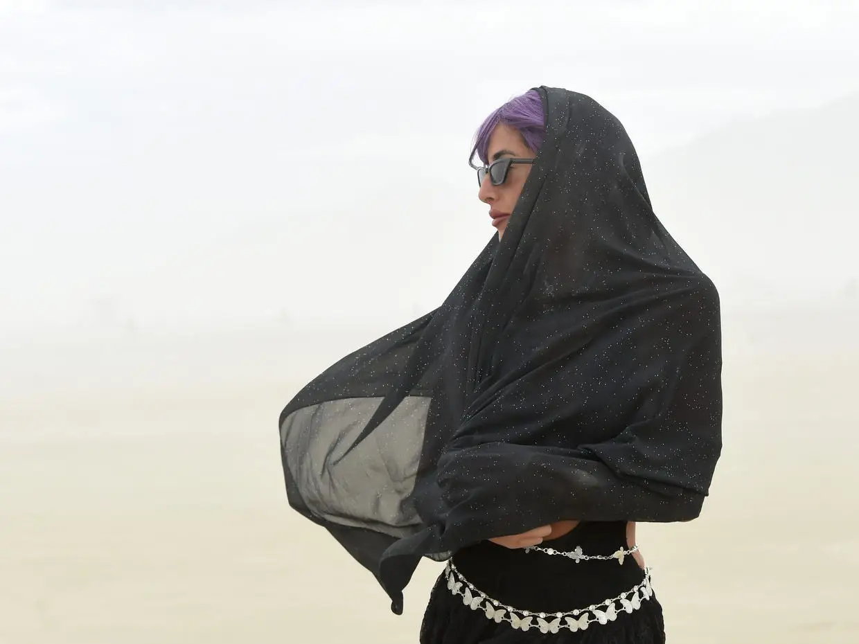 Person with purple hair and sunglasses wears a black sheer scarf in a hazy outdoor setting.
