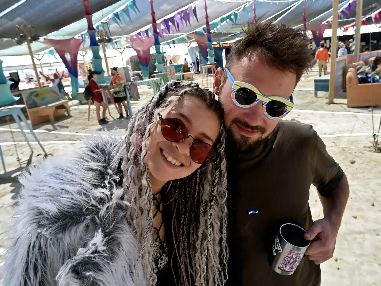 Two festival attendees wearing sunglasses pose under a decorated tent.