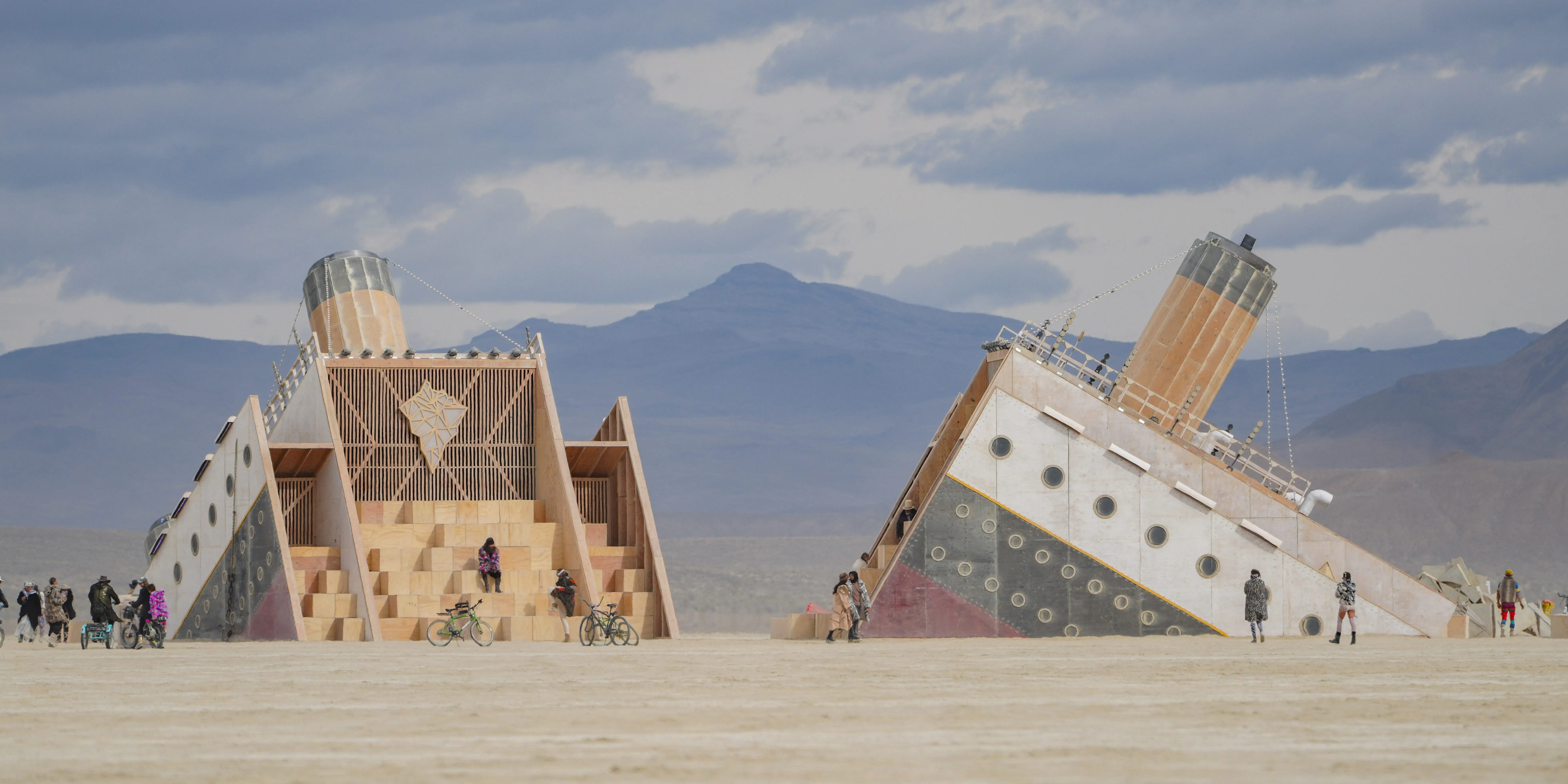 Two large wooden desert structures stand on a flat playa with people and bicycles under a cloudy sky.
