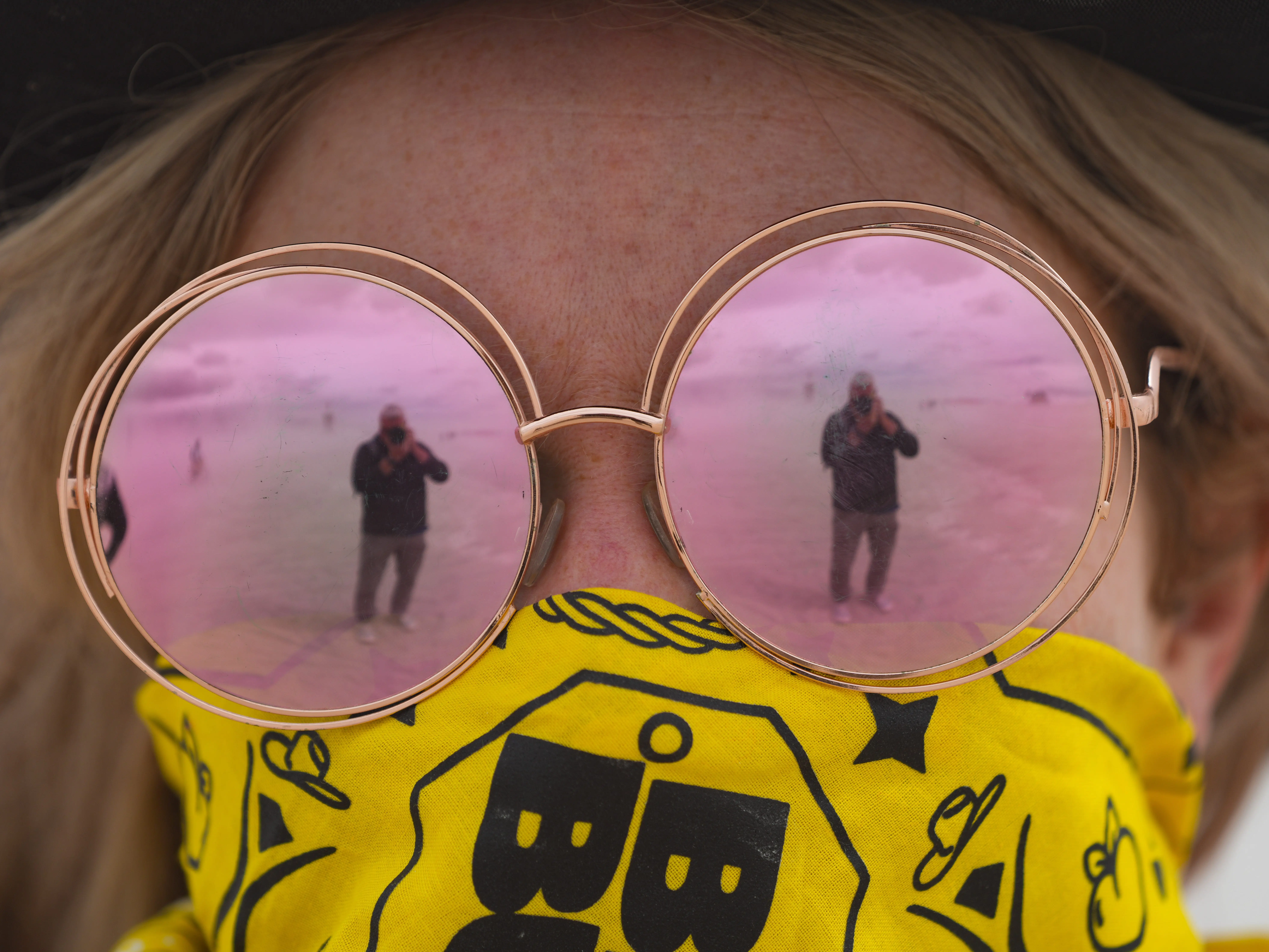 Close-up person wearing round pink reflective sunglasses and a yellow patterned face.