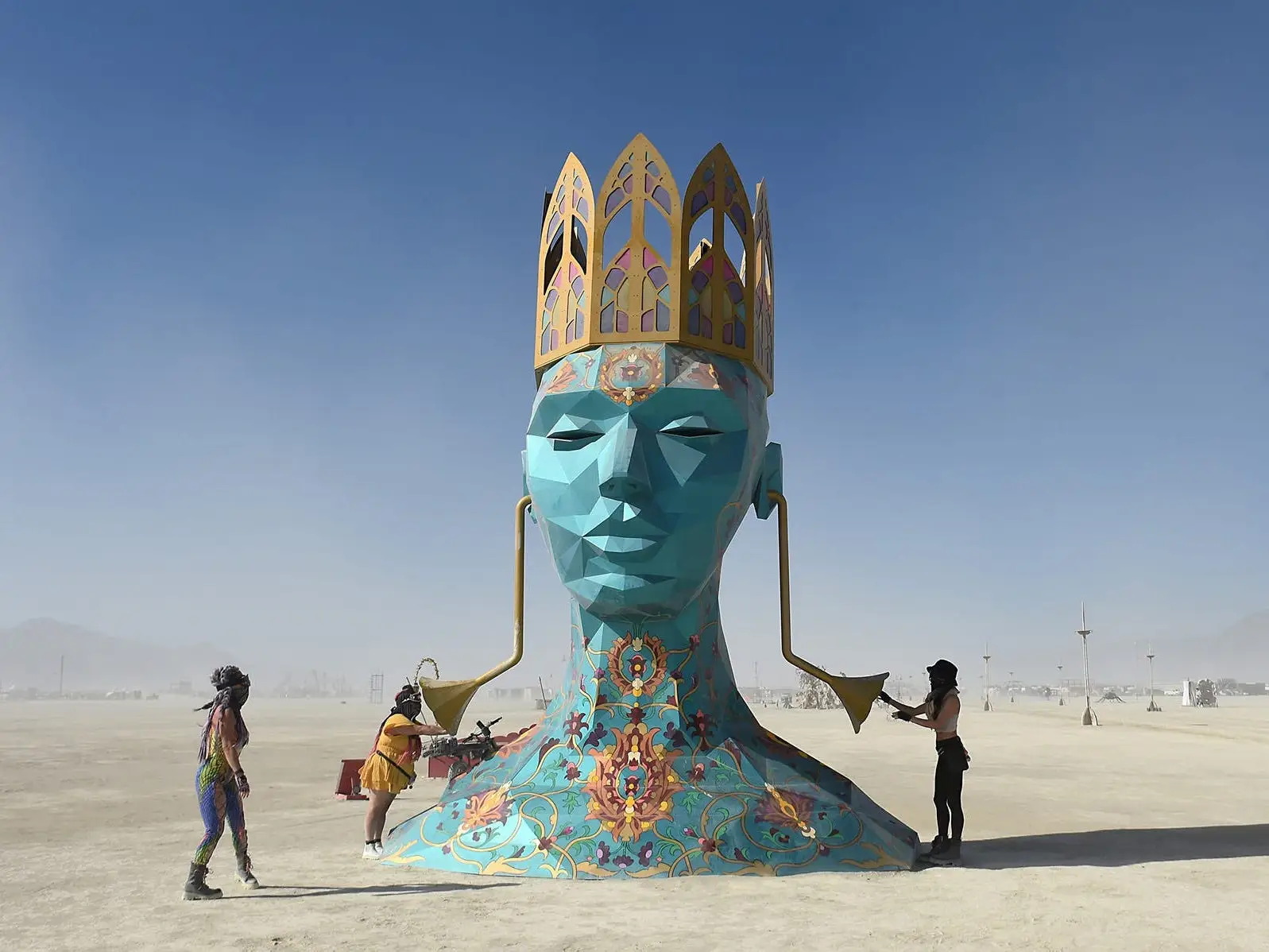 Large blue crowned head sculpture stands on a desert playa with visitors.