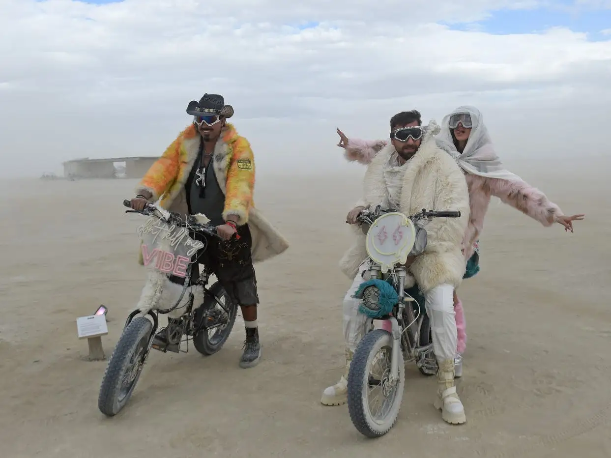 Three costumed riders pose with decorated electric bikes in a dusty desert setting under a cloudy sky.