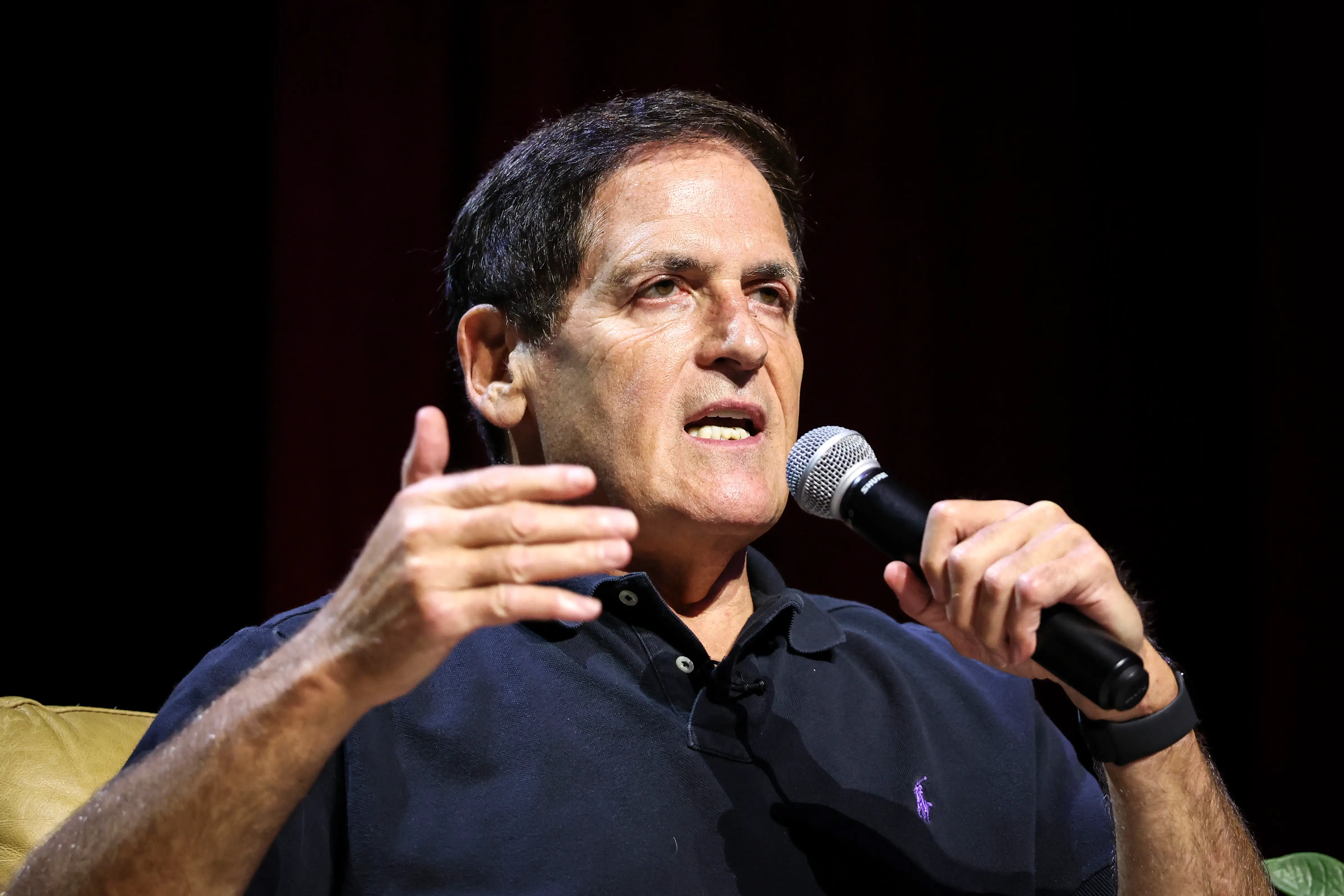 Mark Cuban speaks into a handheld microphone during a stage discussion against a dark backdrop.