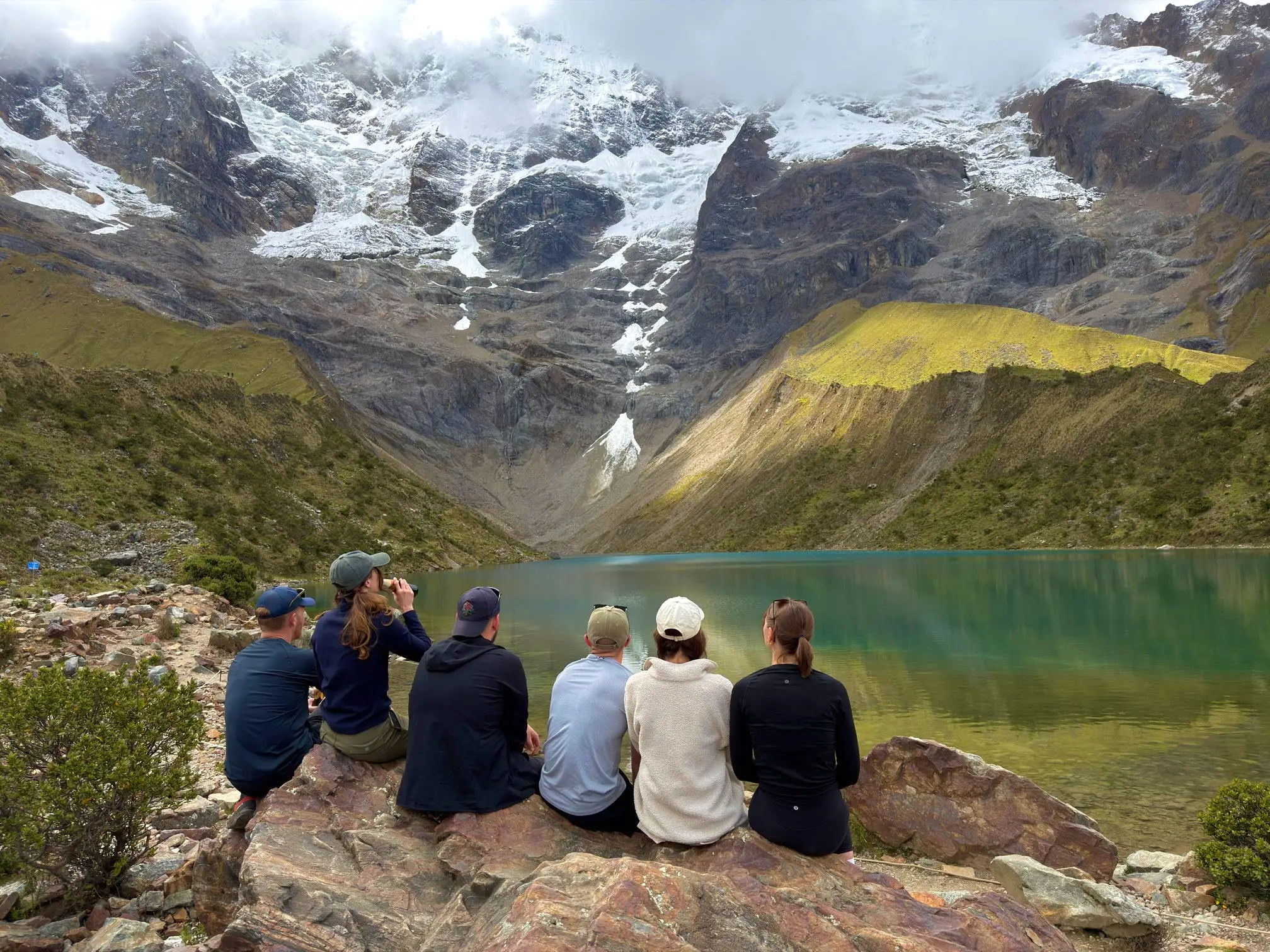 A group of hikers sit on a rock with a snow-capped mountain in the background.