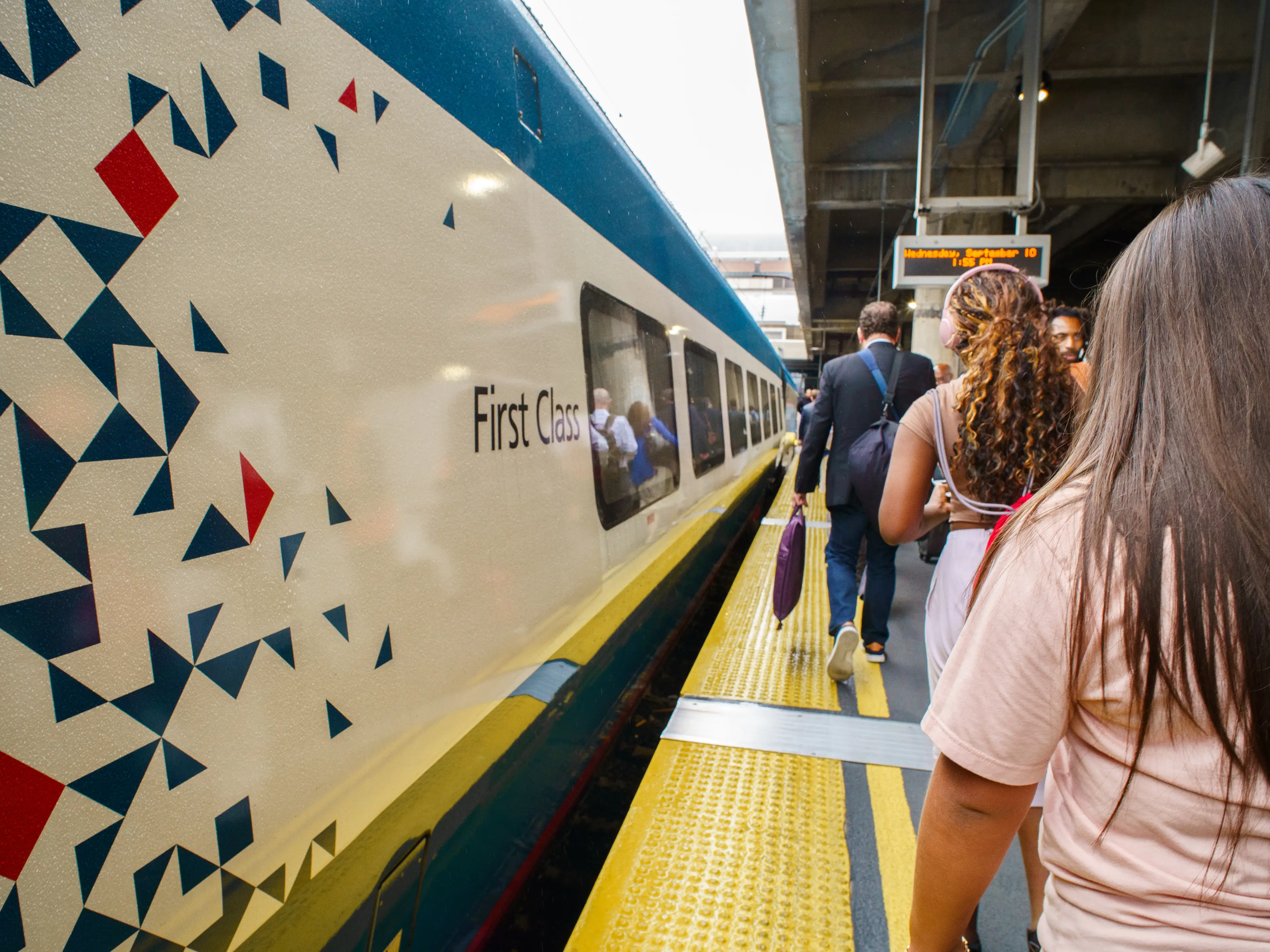 Passengers wait beside an Amtrak NextGen Acela train car marked First Class on a station platform.