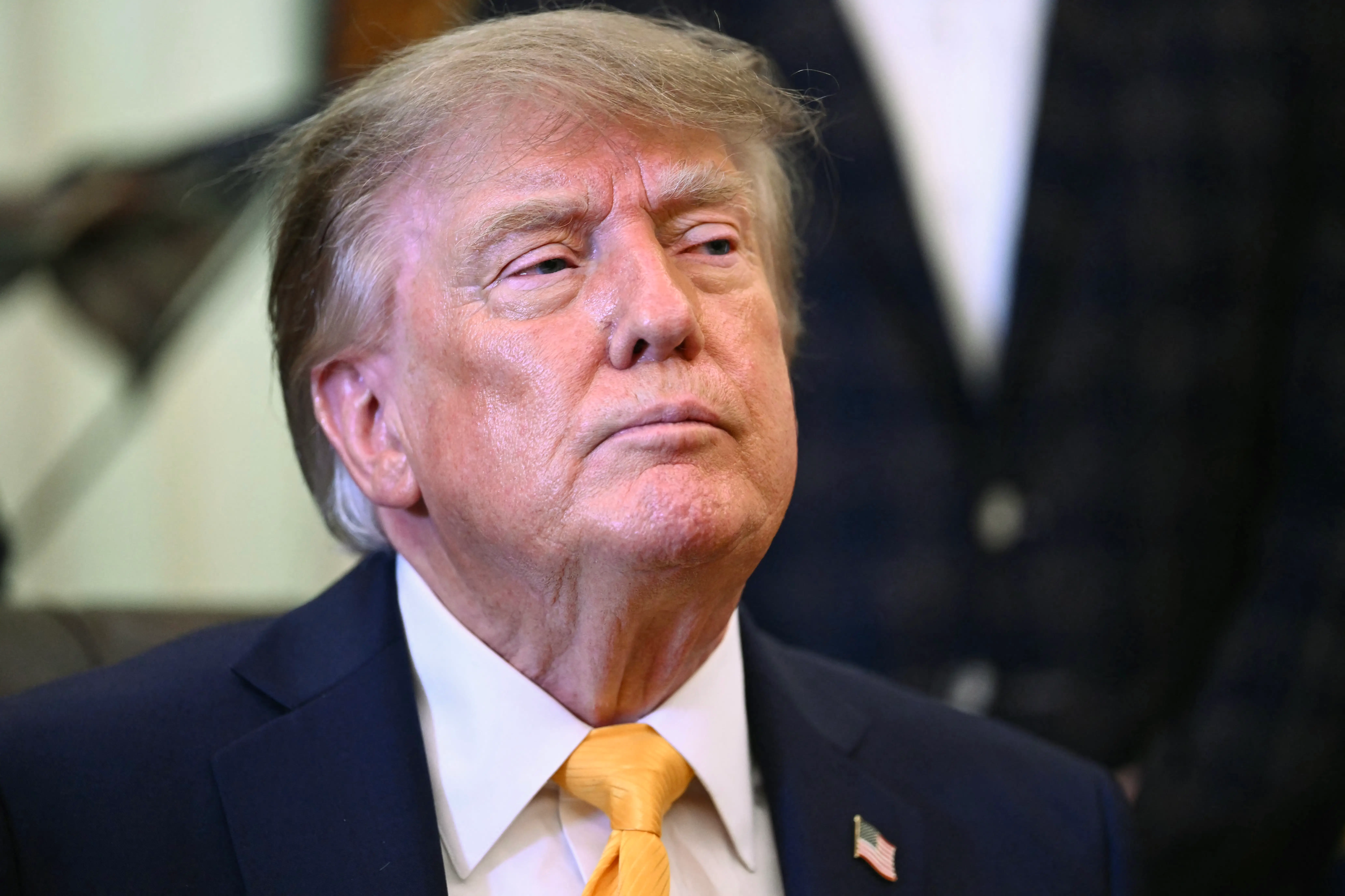 Donald Trump sits in a dark suit with a yellow tie and U.S. flag lapel pin.