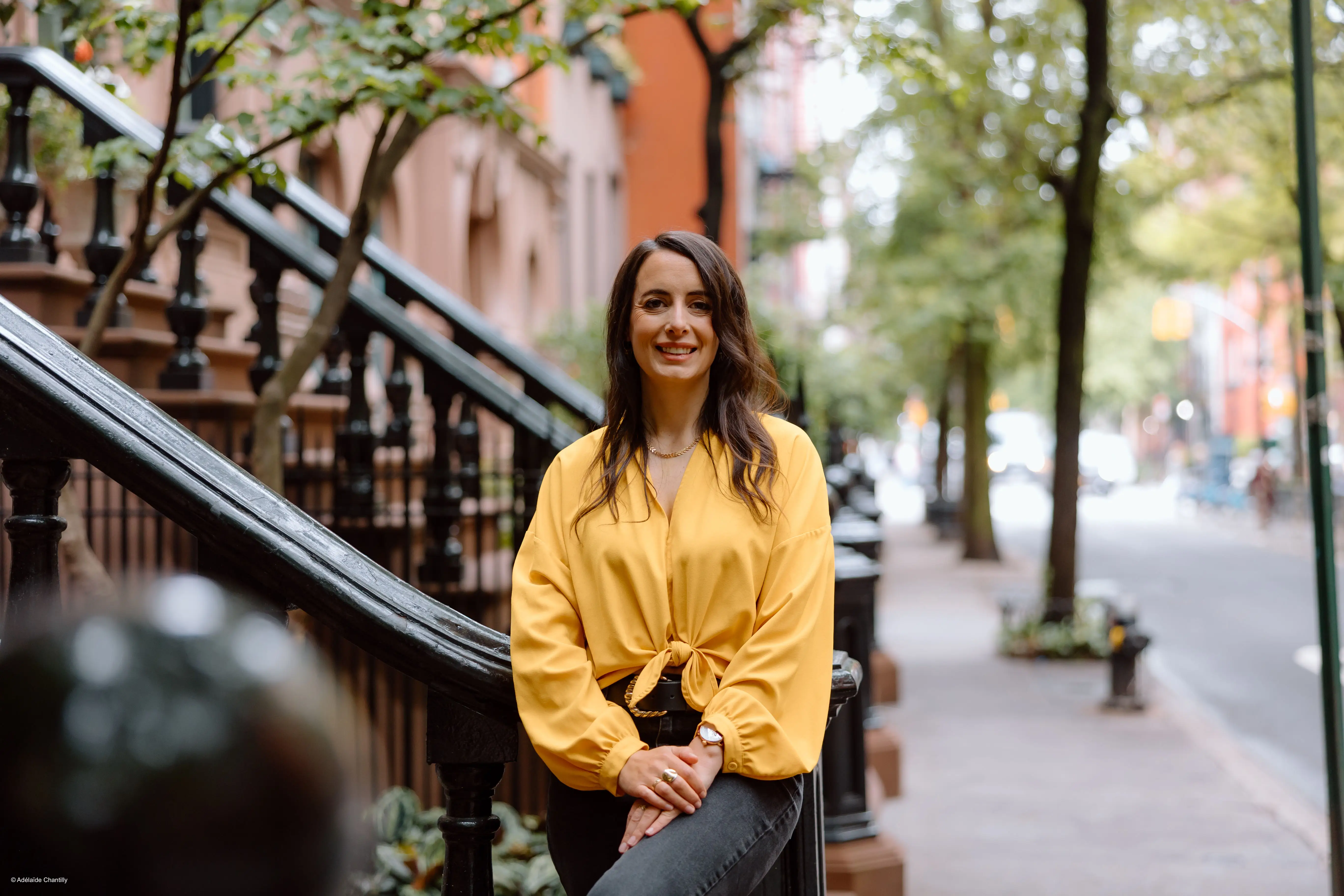 Julie Chapon in a yellow blouse sits on a brownstone stoop along a tree-lined city sidewalk.