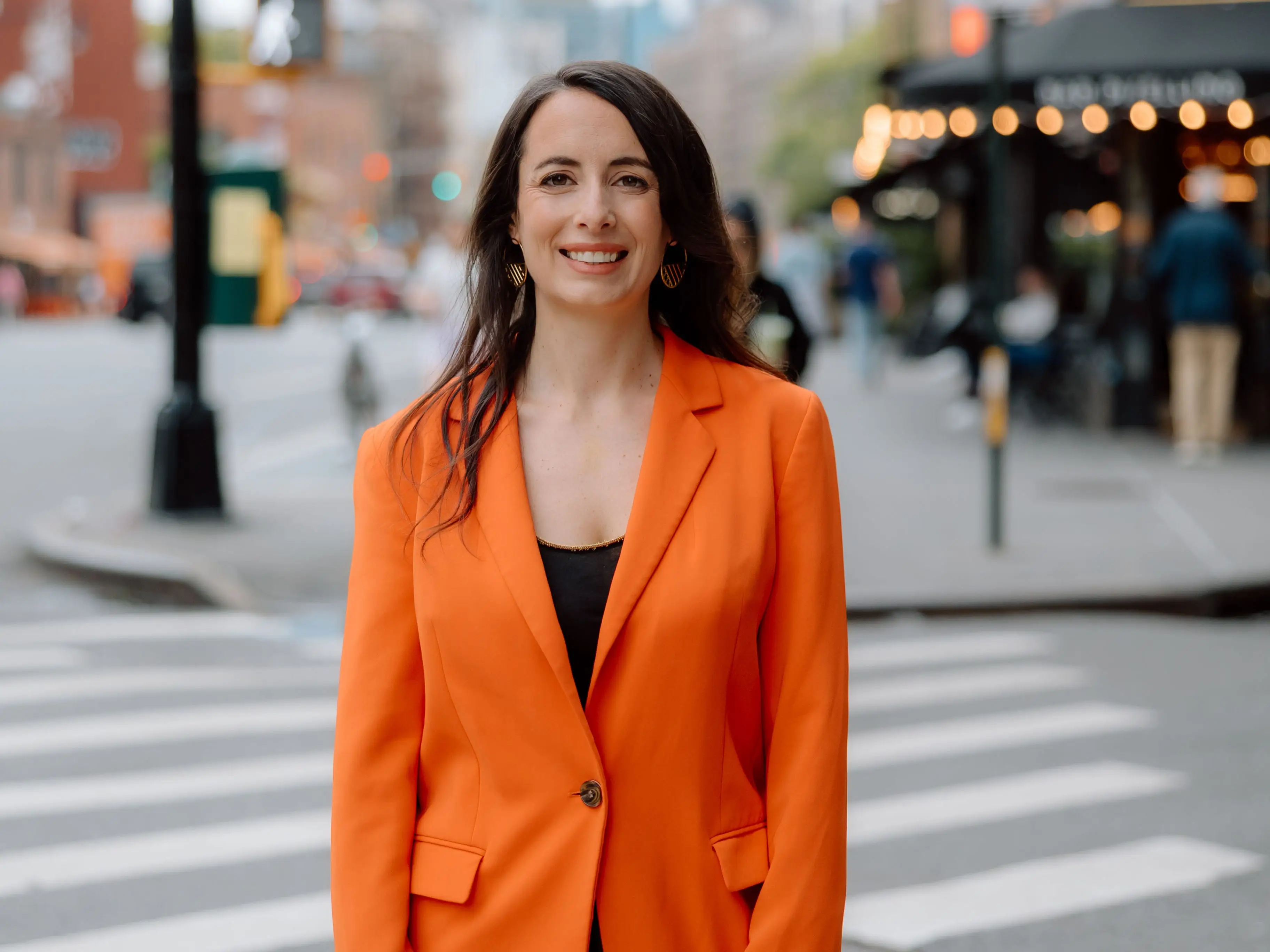 Julie Chapon in a bright orange blazer stands on a city crosswalk with blurred storefronts behind.