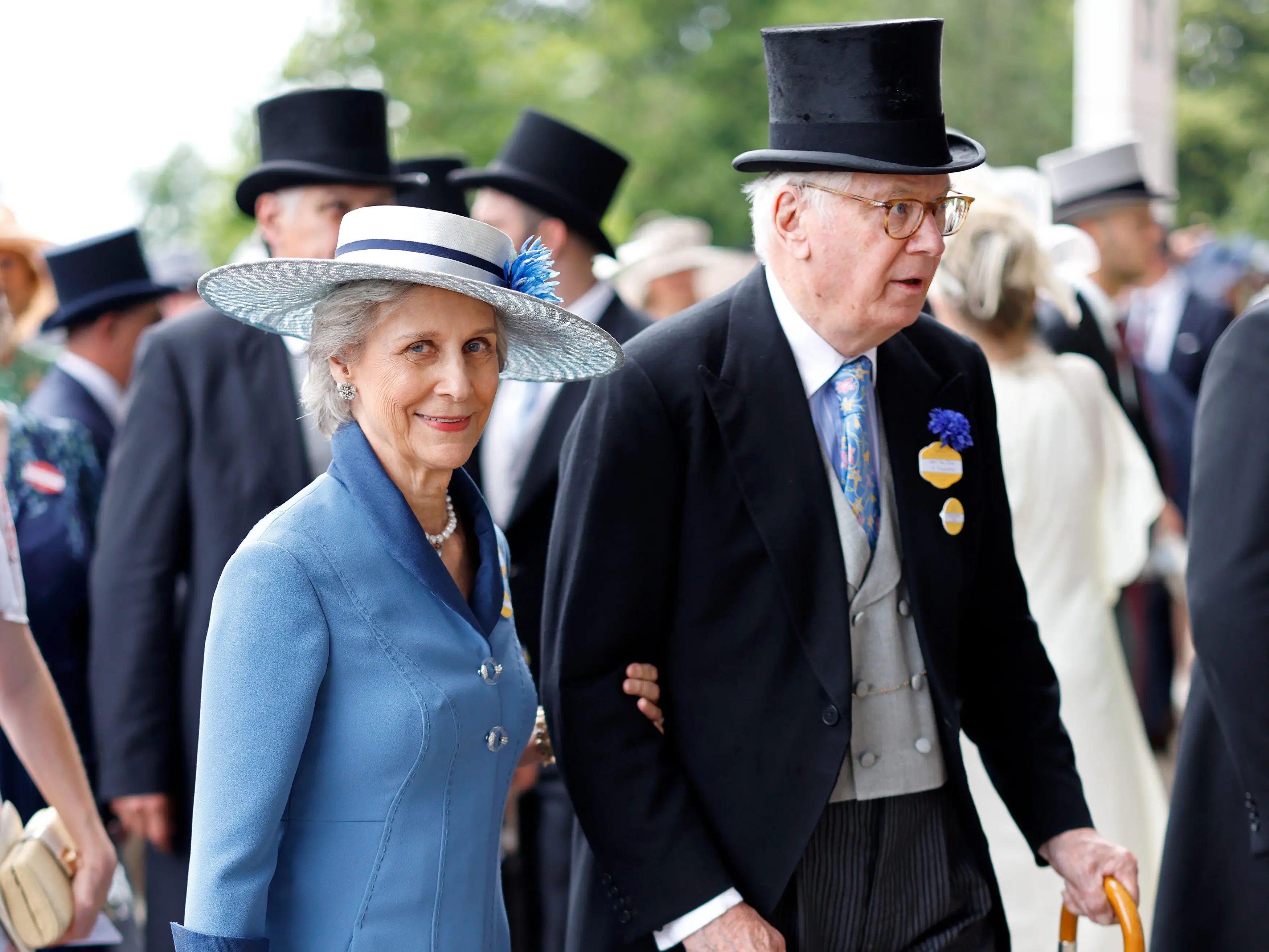 Two formally dressed attendees walk arm in arm at an outdoor event among top-hatted guests.