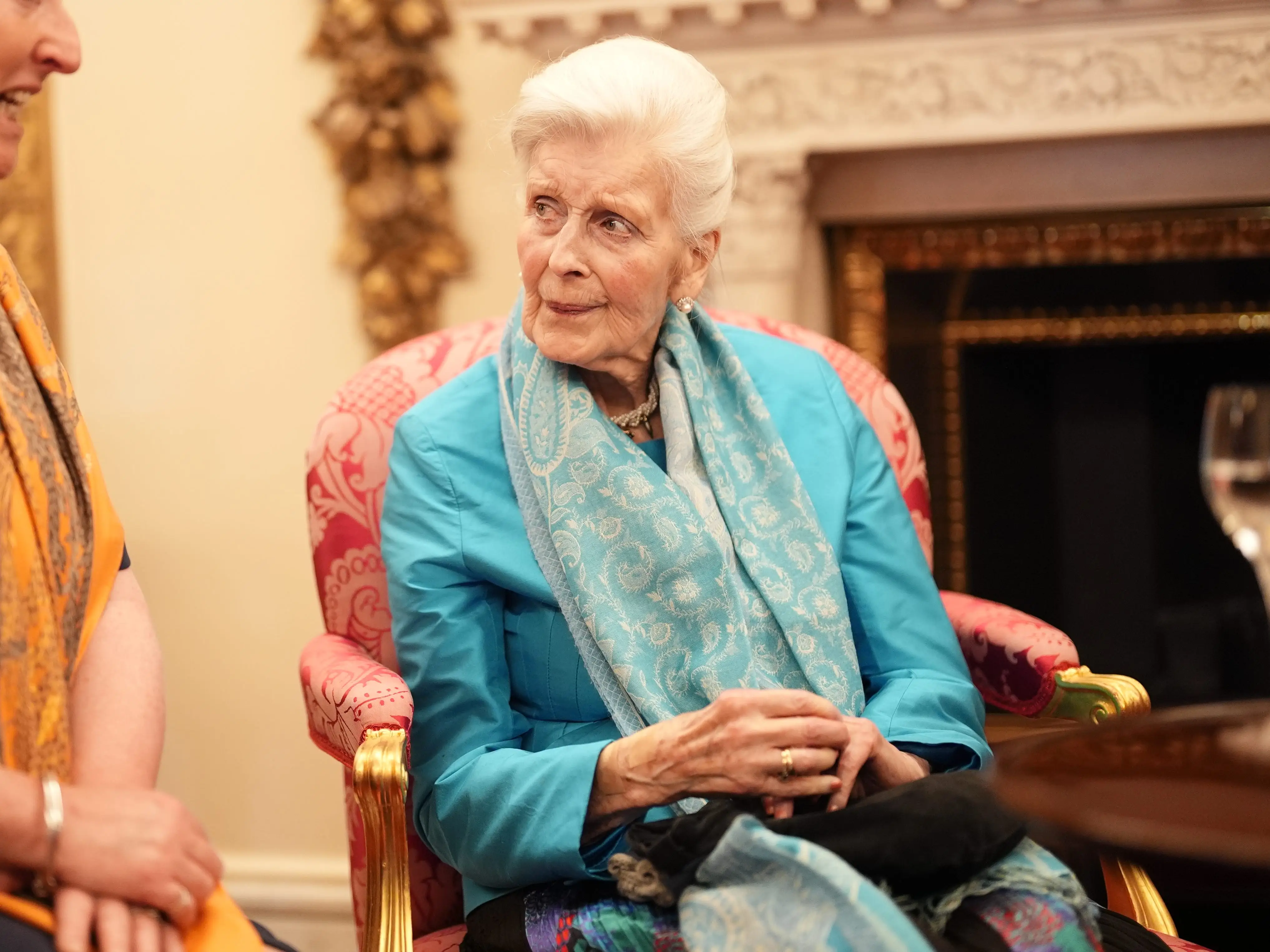 Two people sit in ornate chairs and converse in a warmly lit formal room with framed art.