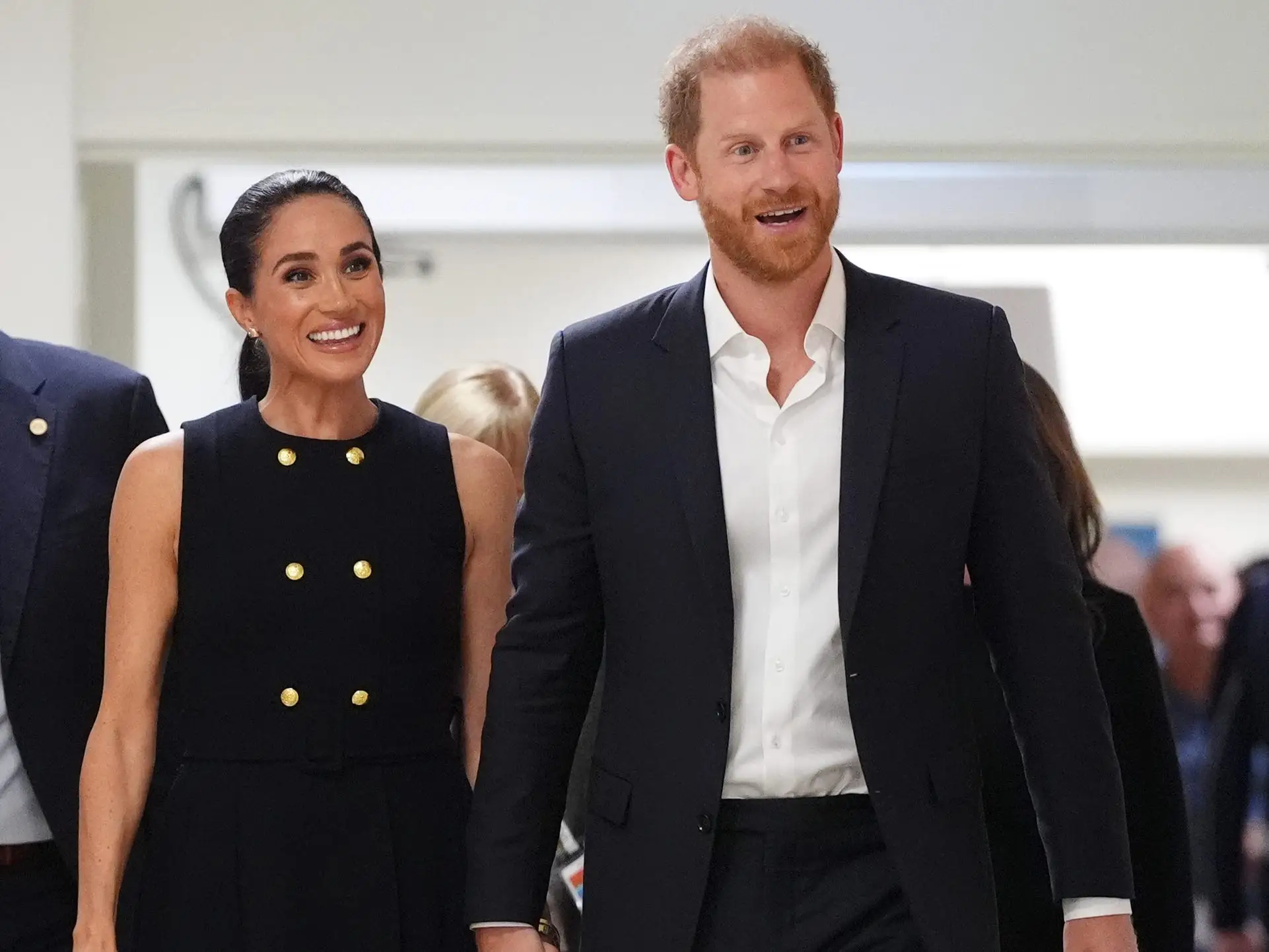 Meghan Markle and Prince Harry walk hand in hand through an indoor corridor in formal black outfits.