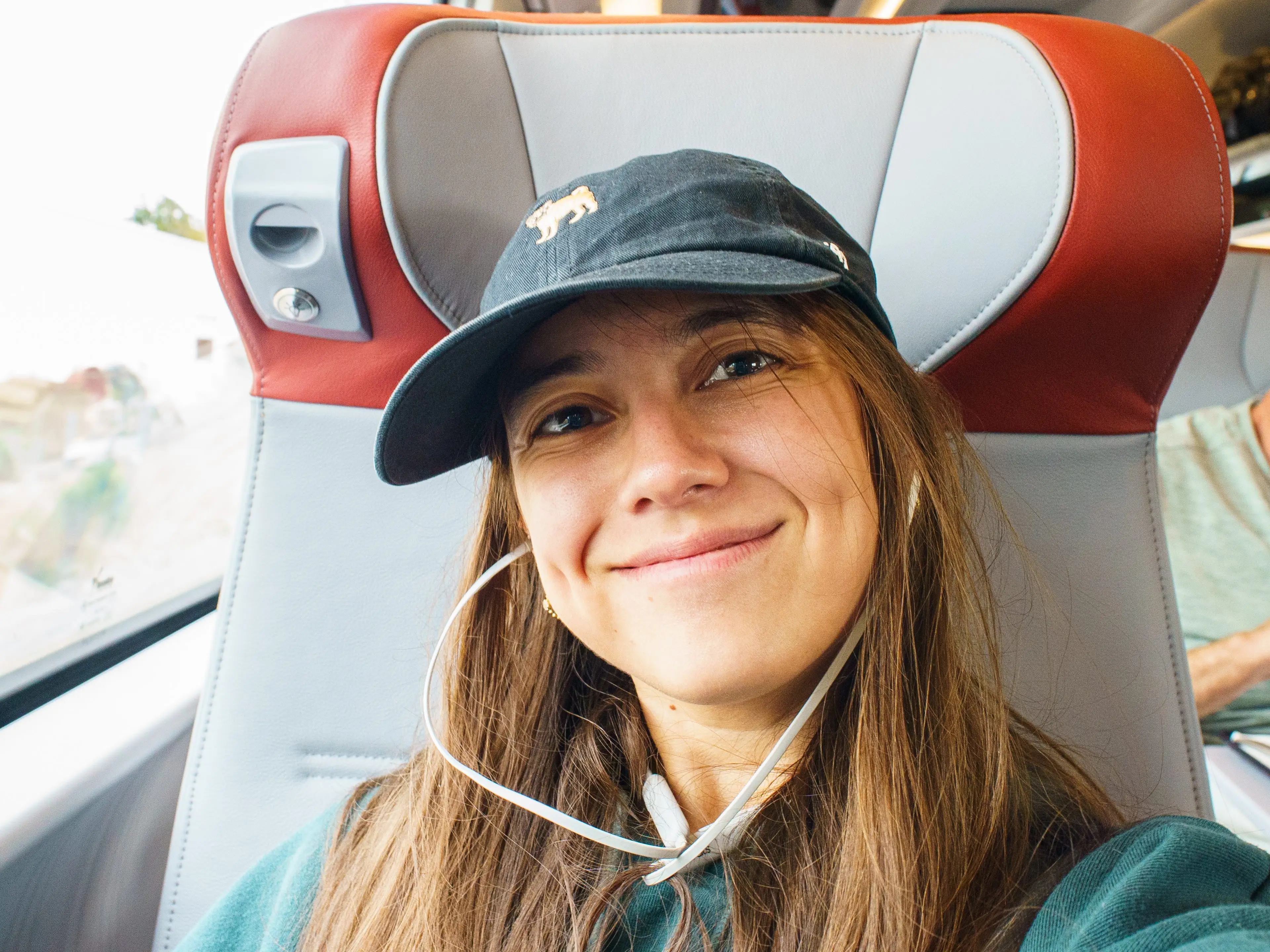Smiling passenger wearing earbuds and a black cap sits in a red-and-gray train seat.
