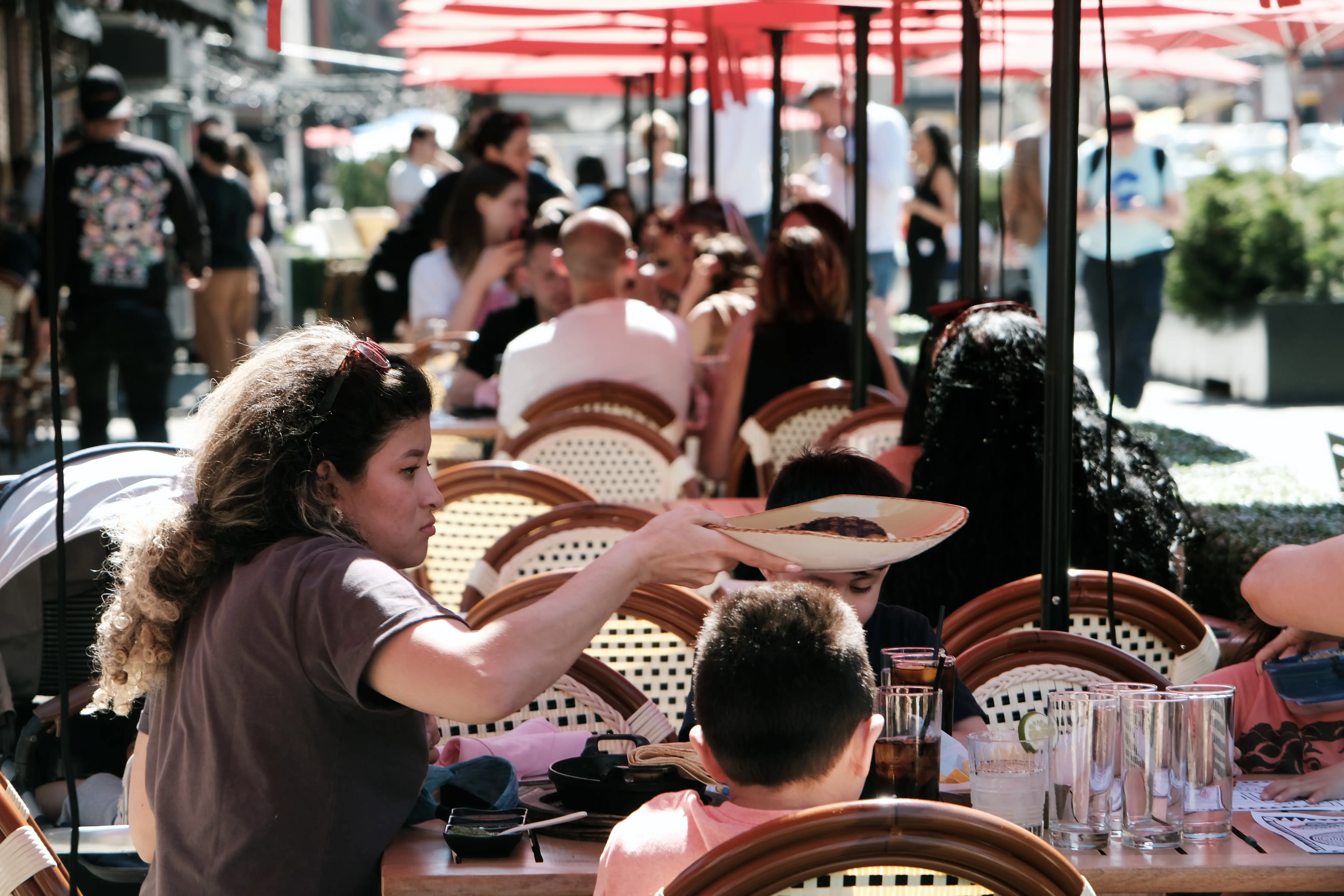 A busy outdoor restaurant patio with red umbrellas, diners, and a person reaching across a table with a plate.