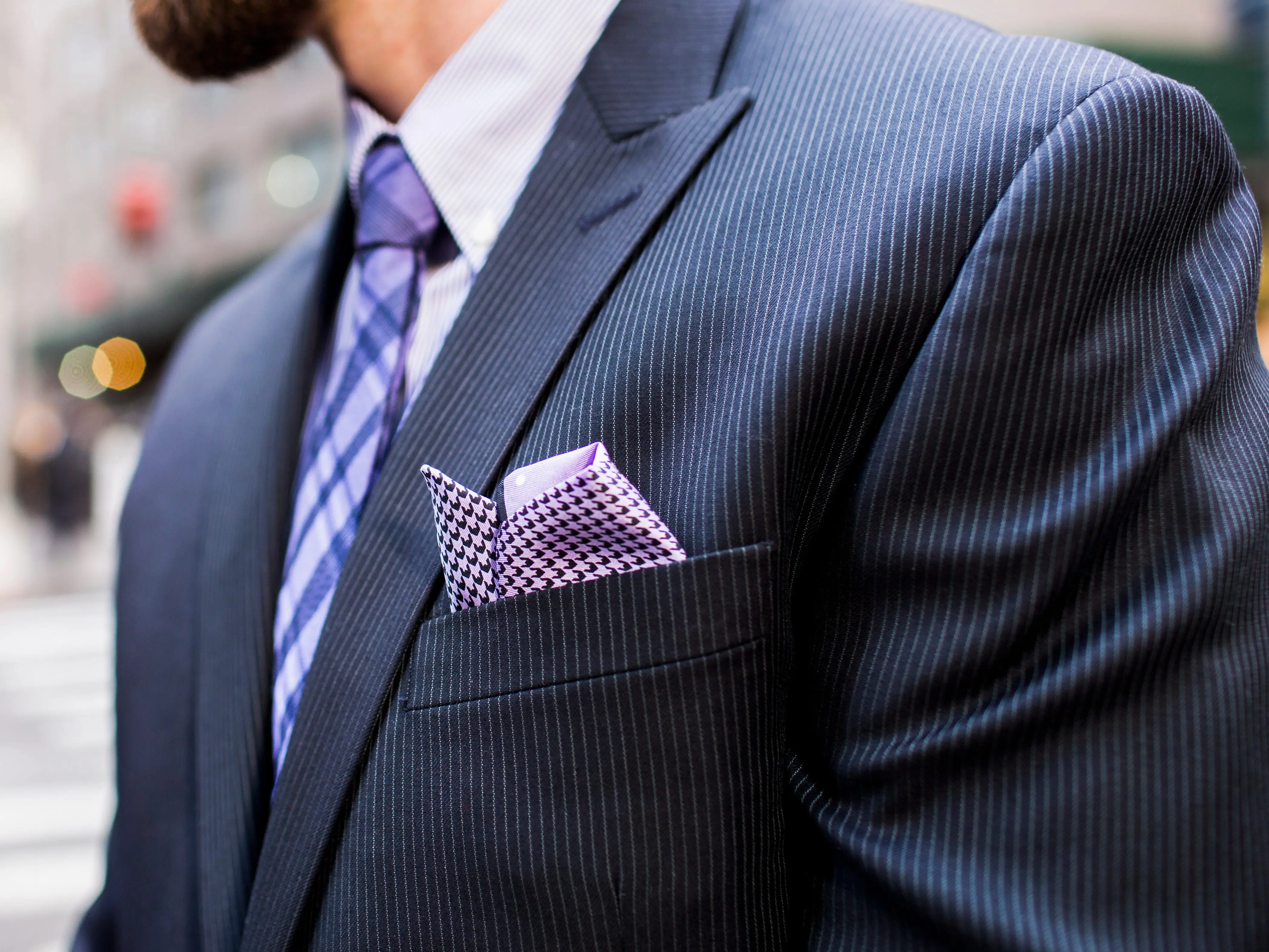 Close-up of a man wearing a pinstripe suit, purple tie, and patterned pocket square on a city street.