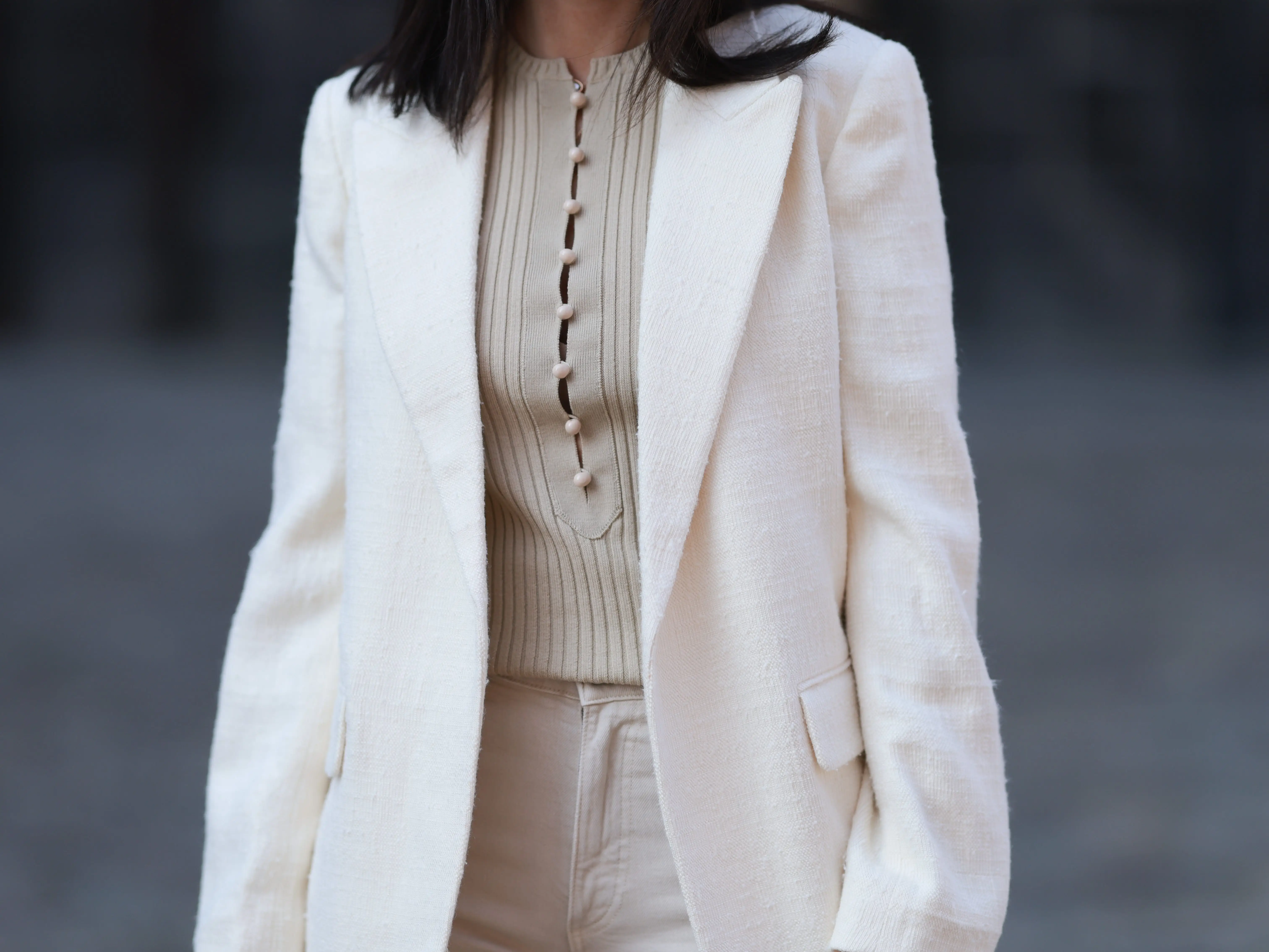 A woman wearing a cream blazer over a ribbed buttoned top and light trousers on a blurred city street.