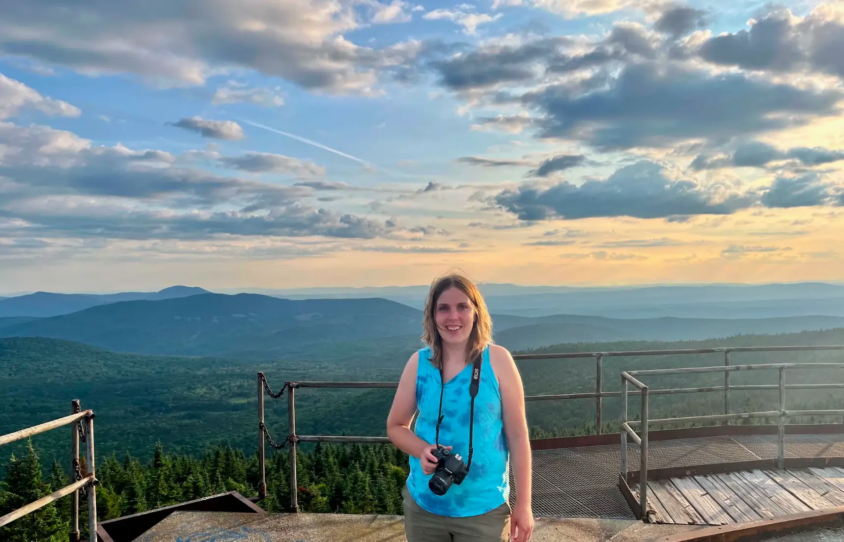 The writer wearing a blue shirt, holding a camera, and standing in front of an autumn landscape in New England.