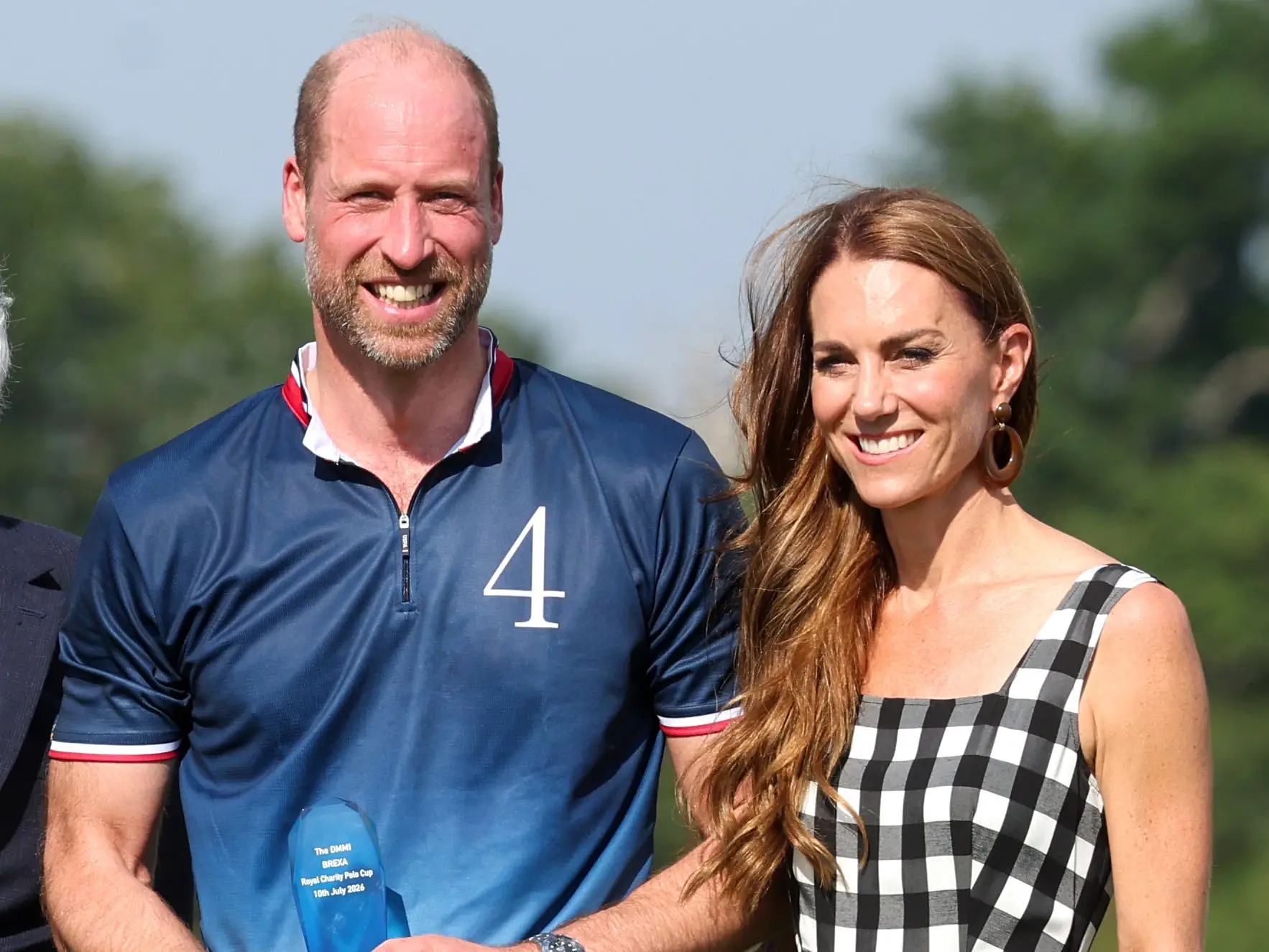 Prince William holds a blue trophy beside Catherine, Princess of Wales, on a sunny polo field.